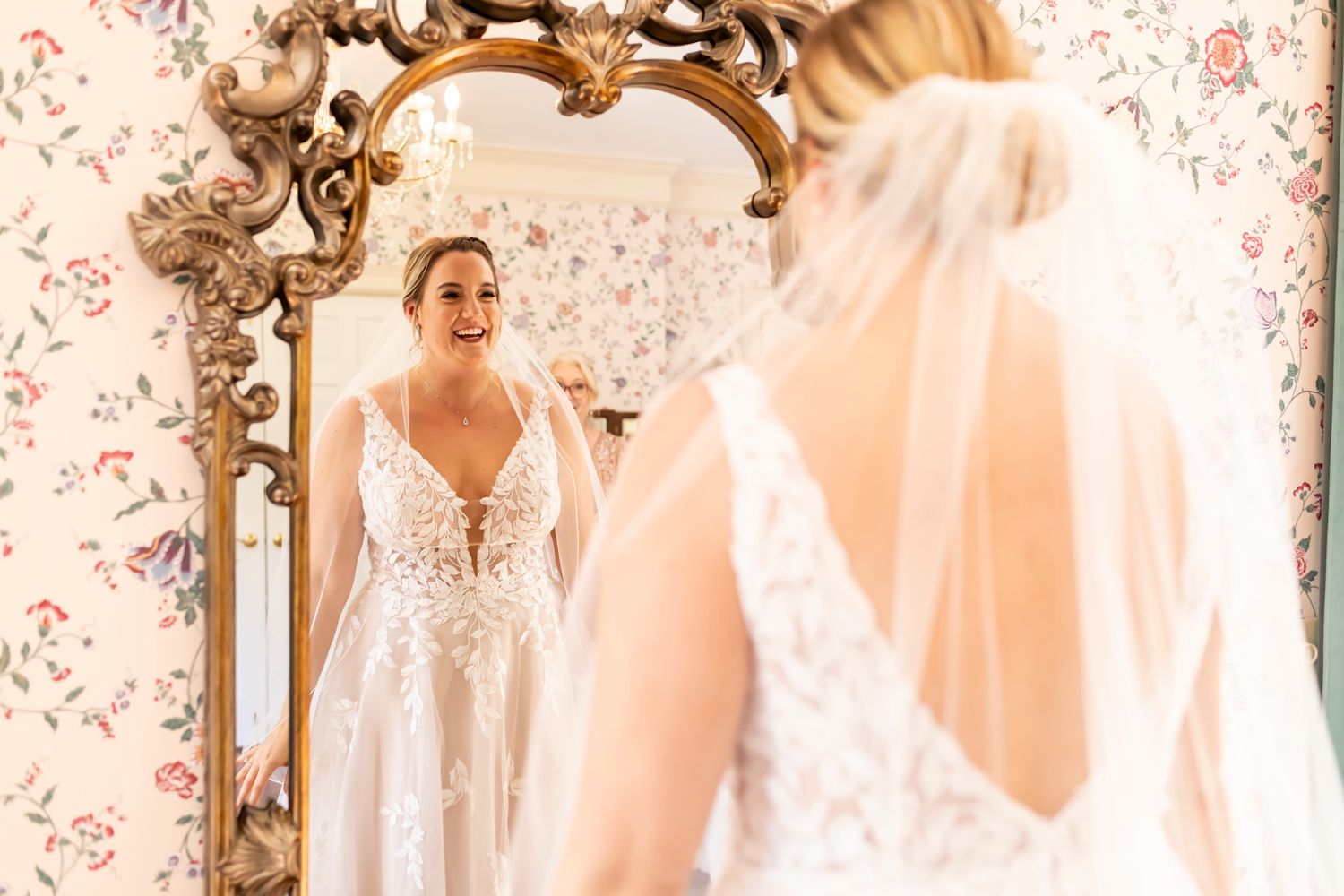 Bride wearing white lace wedding dress with plunging neckline gazes at reflection in ornate gold vintage mirror.