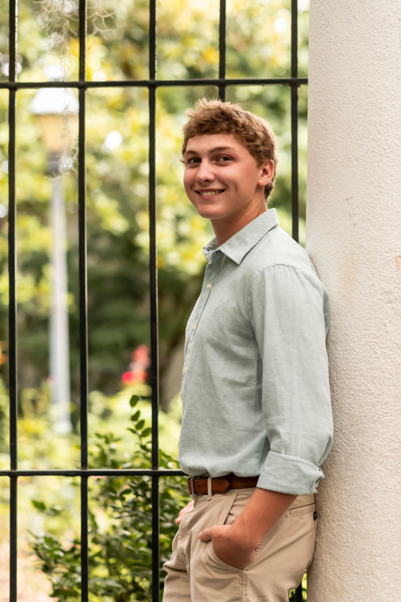 Young person in light dress shirt and khakis poses casually against white column with decorative iron railing.