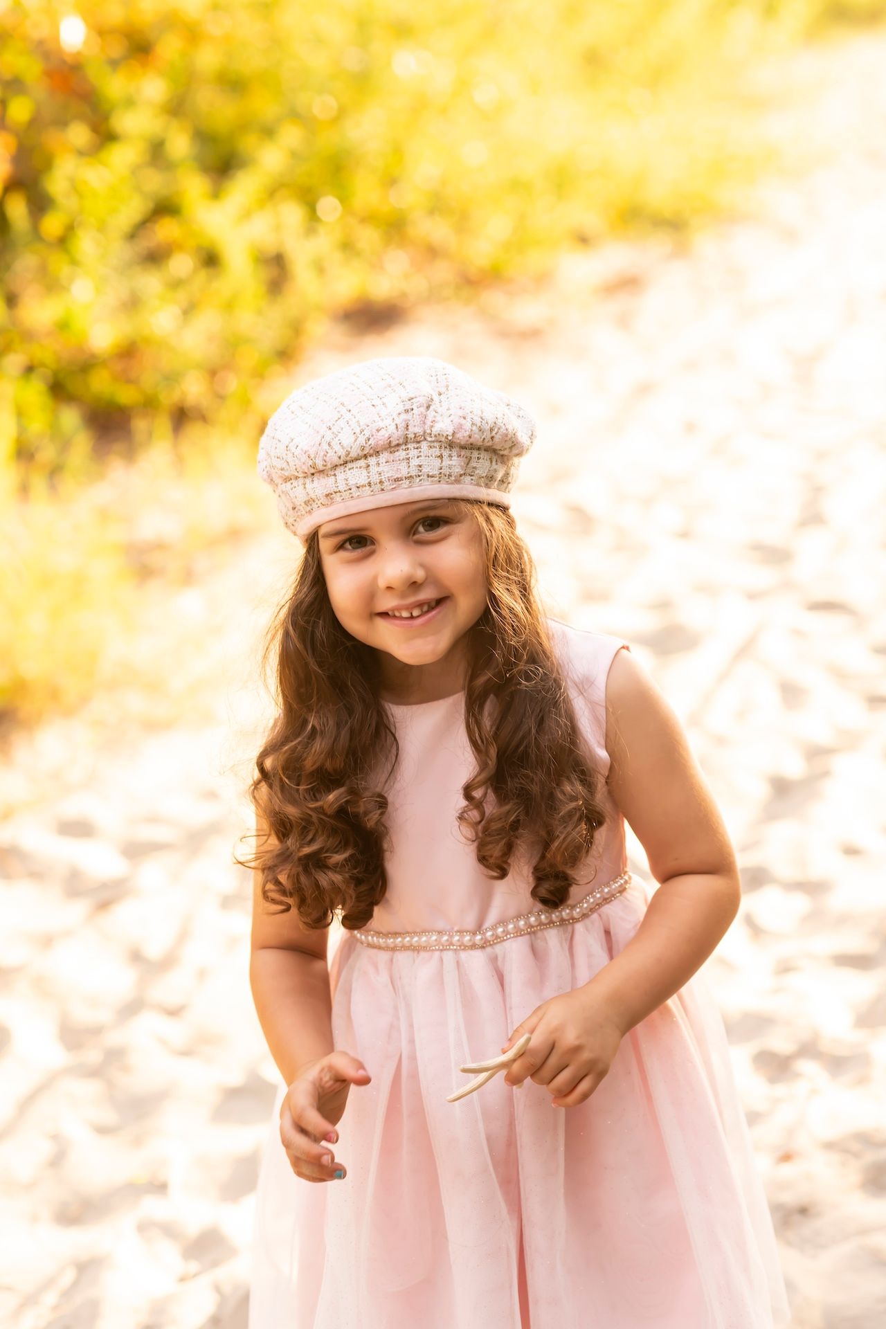 A young child in a pink dress and knit cap poses outdoors against a warm golden autumn background.