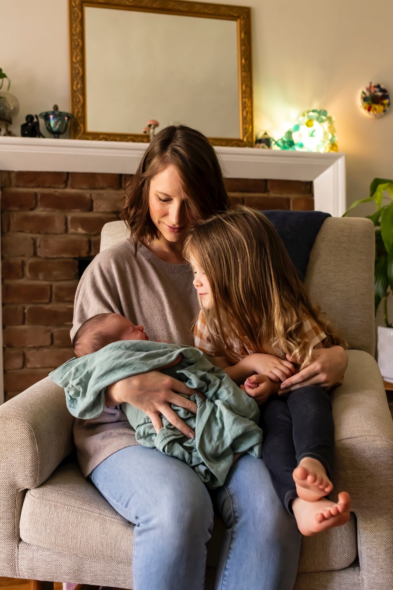 Two siblings sitting in a cozy armchair by a brick fireplace while lovingly looking at a sleeping baby wrapped in a blanket.