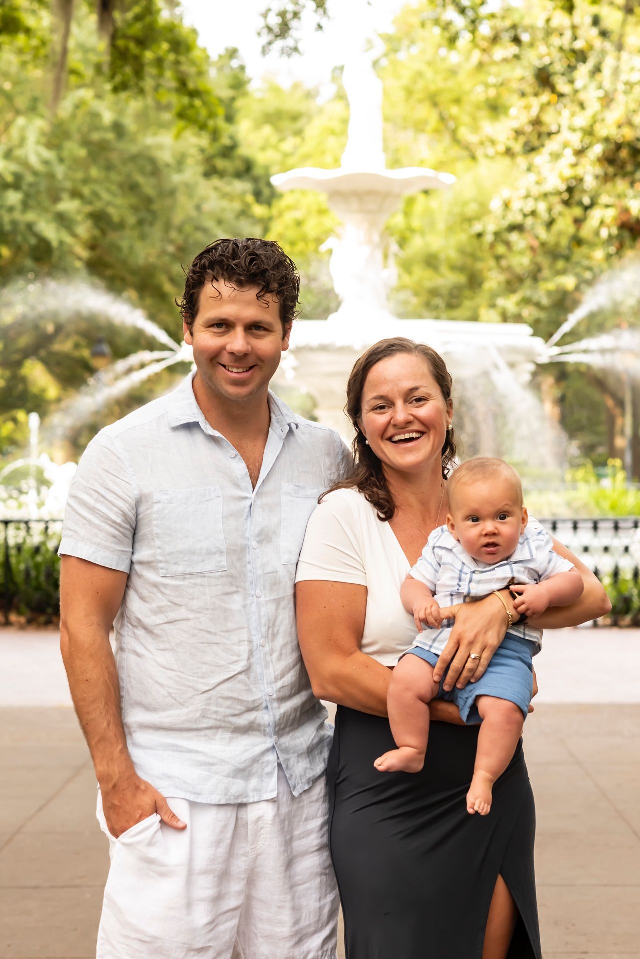 A candid family moment with parents and baby outdoors near a fountain on a bright summer day.