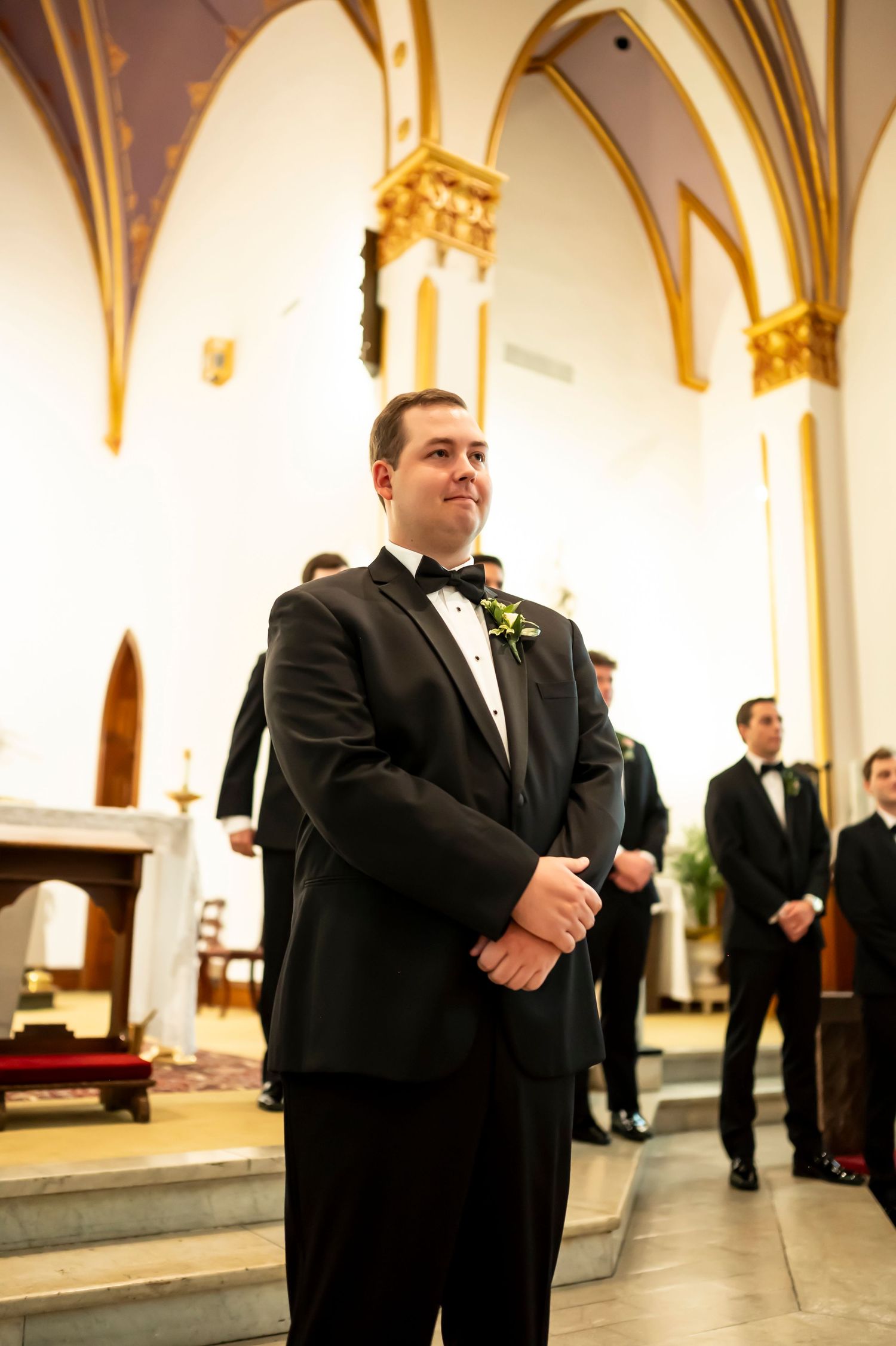 A groom in a classic black tuxedo and bow tie stands at the altar in an ornate church with vaulted gothic arches.