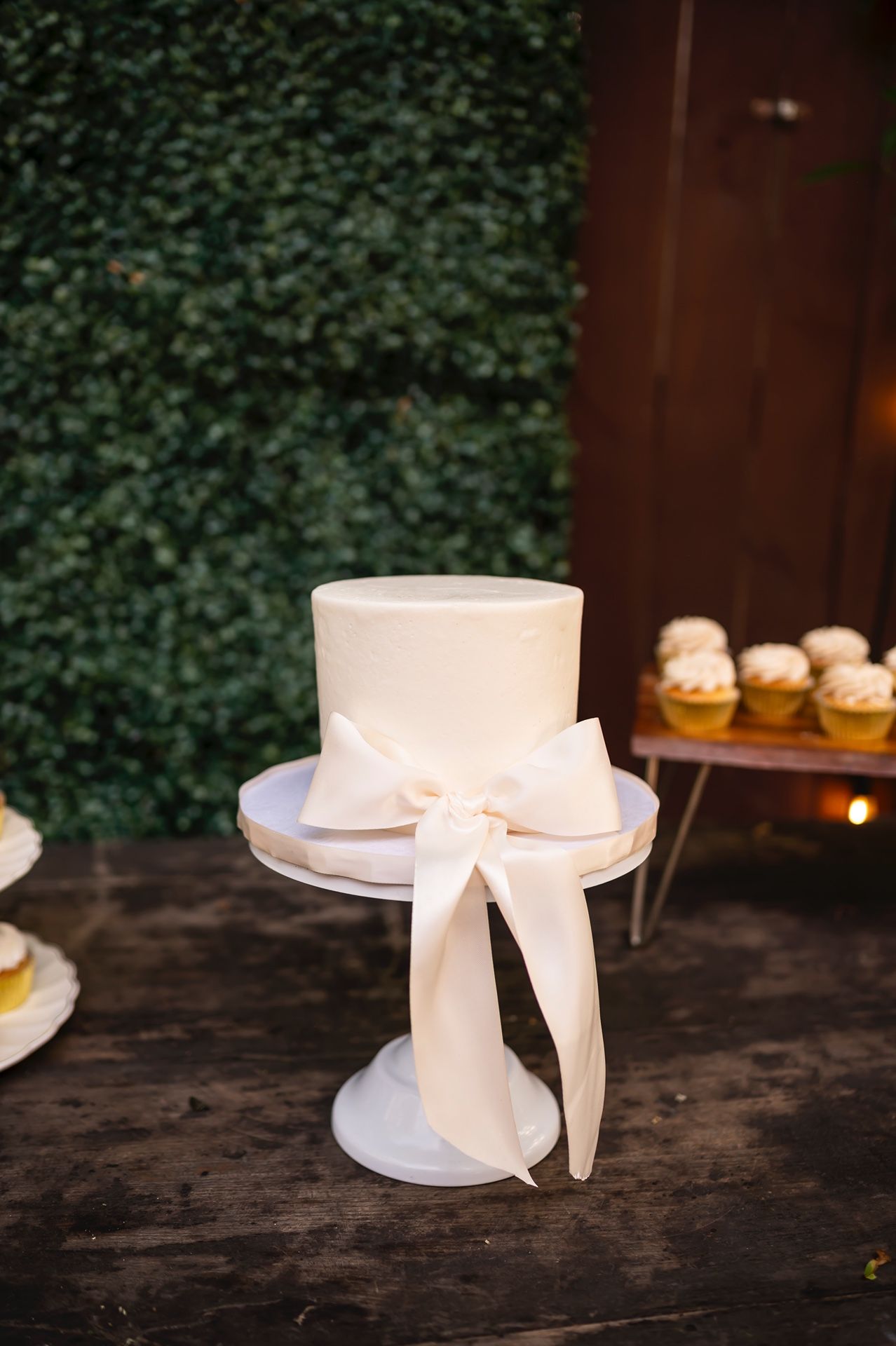 Simple white wedding cake with elegant silk bow ribbon displayed on white pedestal against greenery background.
