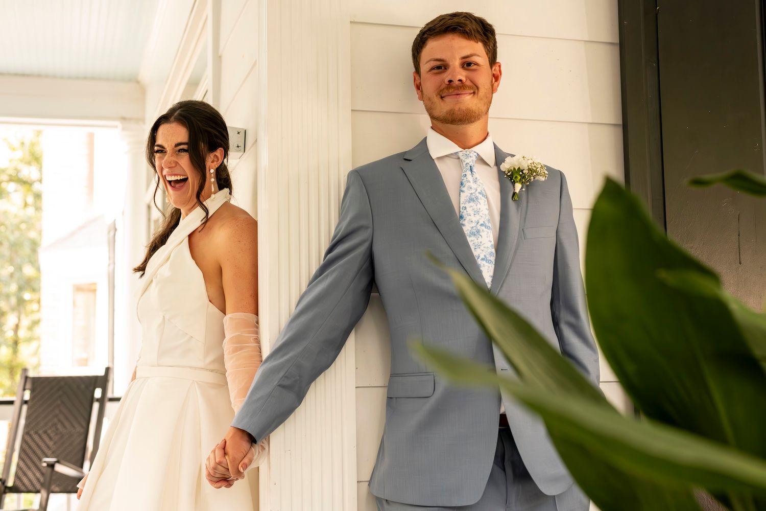 A couple in wedding attire hold hands in a bright indoor setting with a green plant leaf in the foreground.