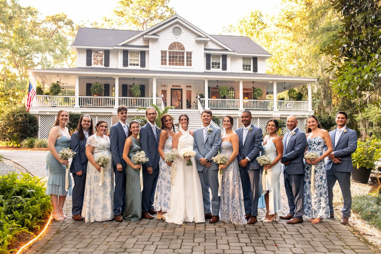 Bridal party standing together on a brick pathway in front of a grand white colonial style house with wrap-around porch.