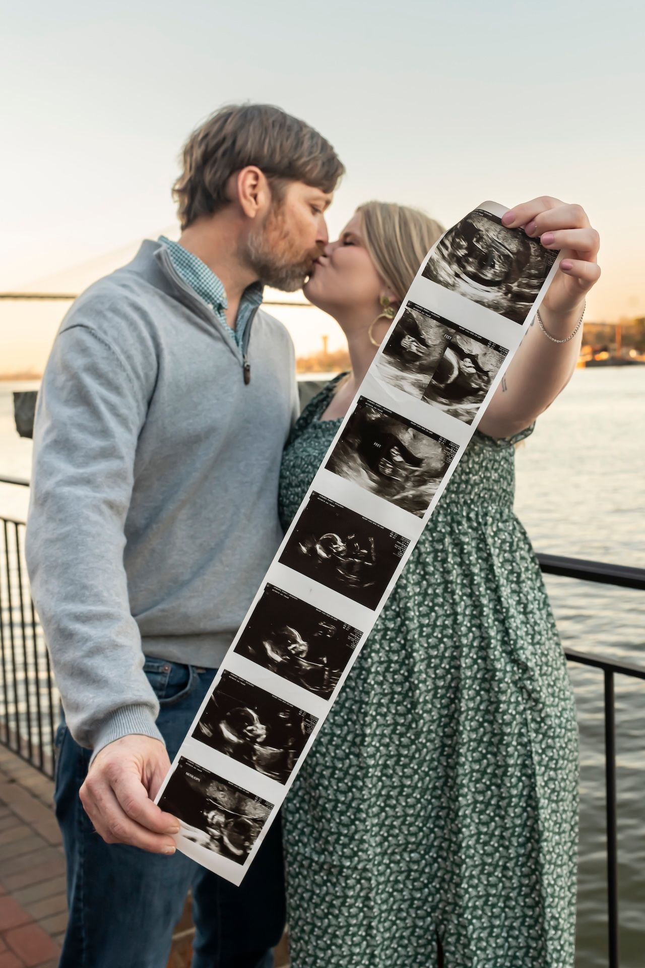 Couple kissing while holding ultrasound photos on waterfront dock during sunset.