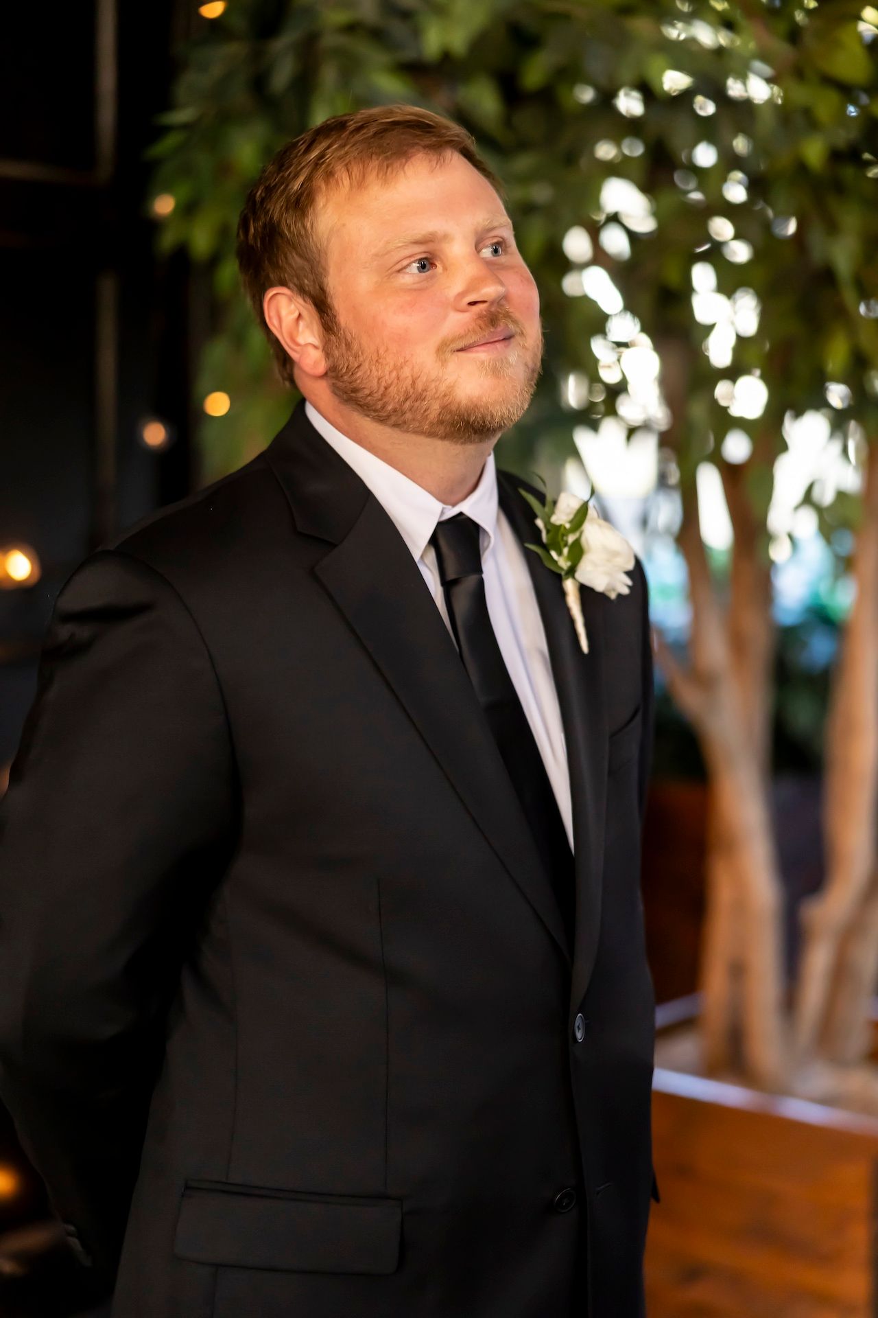A groom in a black tuxedo and tie smiles warmly during a wedding celebration with twinkle lights in the background.