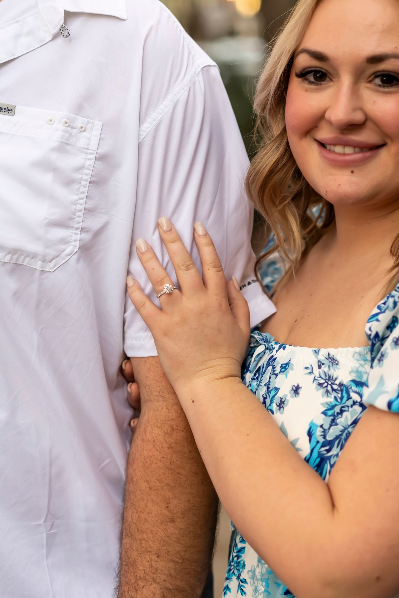 Close up detail of an engagement ring being shown on hand resting against white shirt, with floral dress in view.