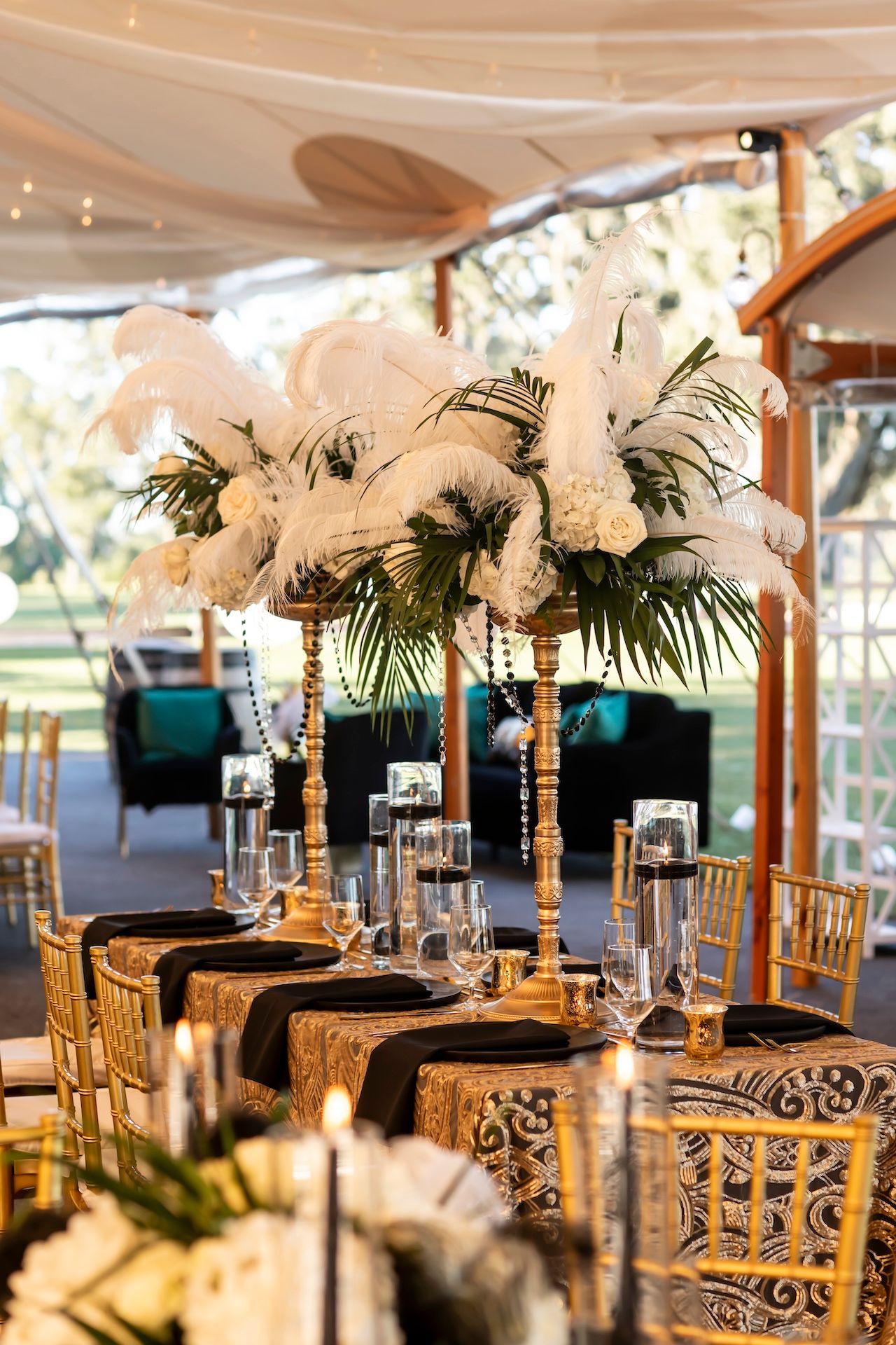 Elegant wedding table setting with tall white feather centerpieces, gold chairs, and black linens under a draped tent ceiling.