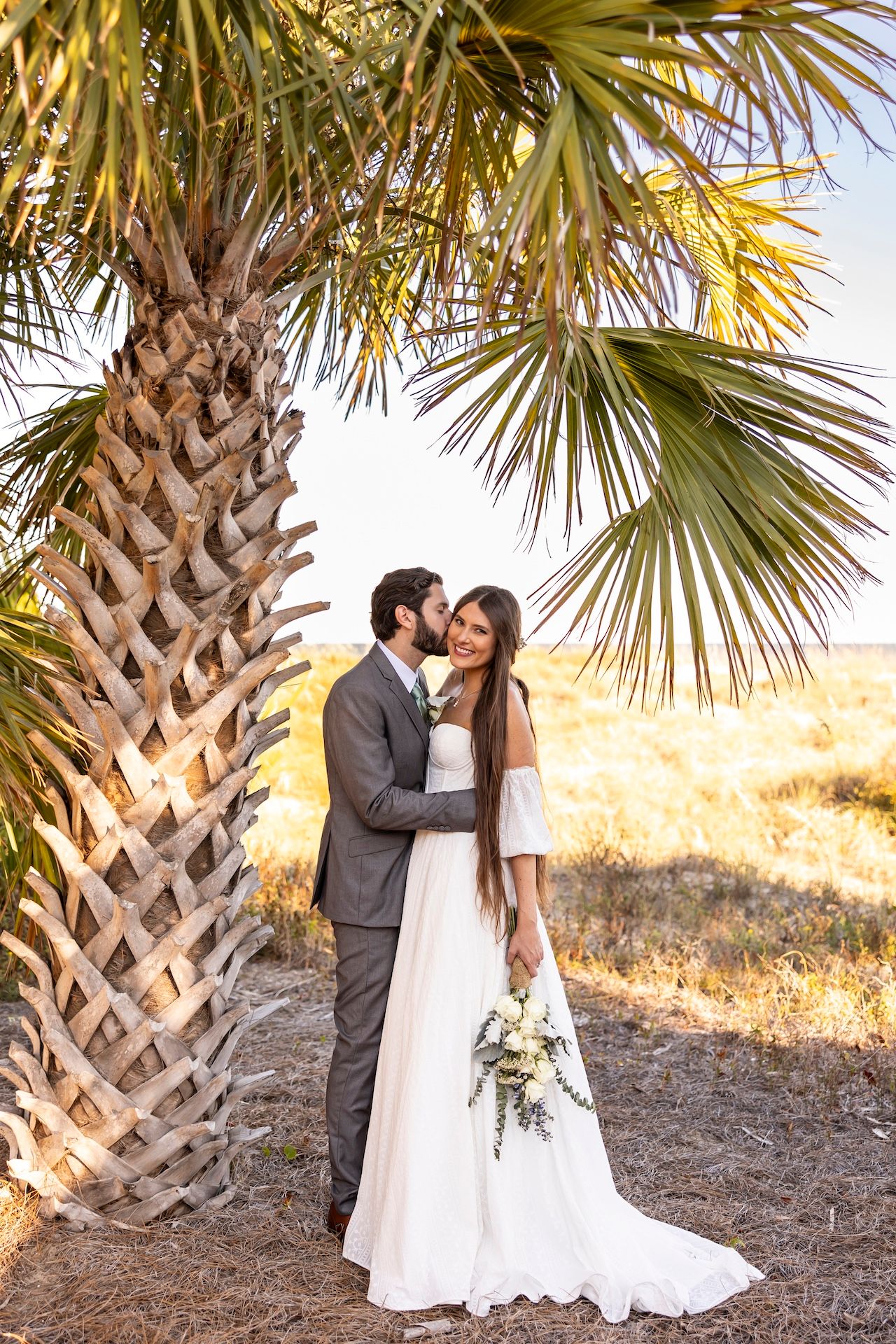 A romantic wedding couple embraces under a palm tree at sunset on a beach.