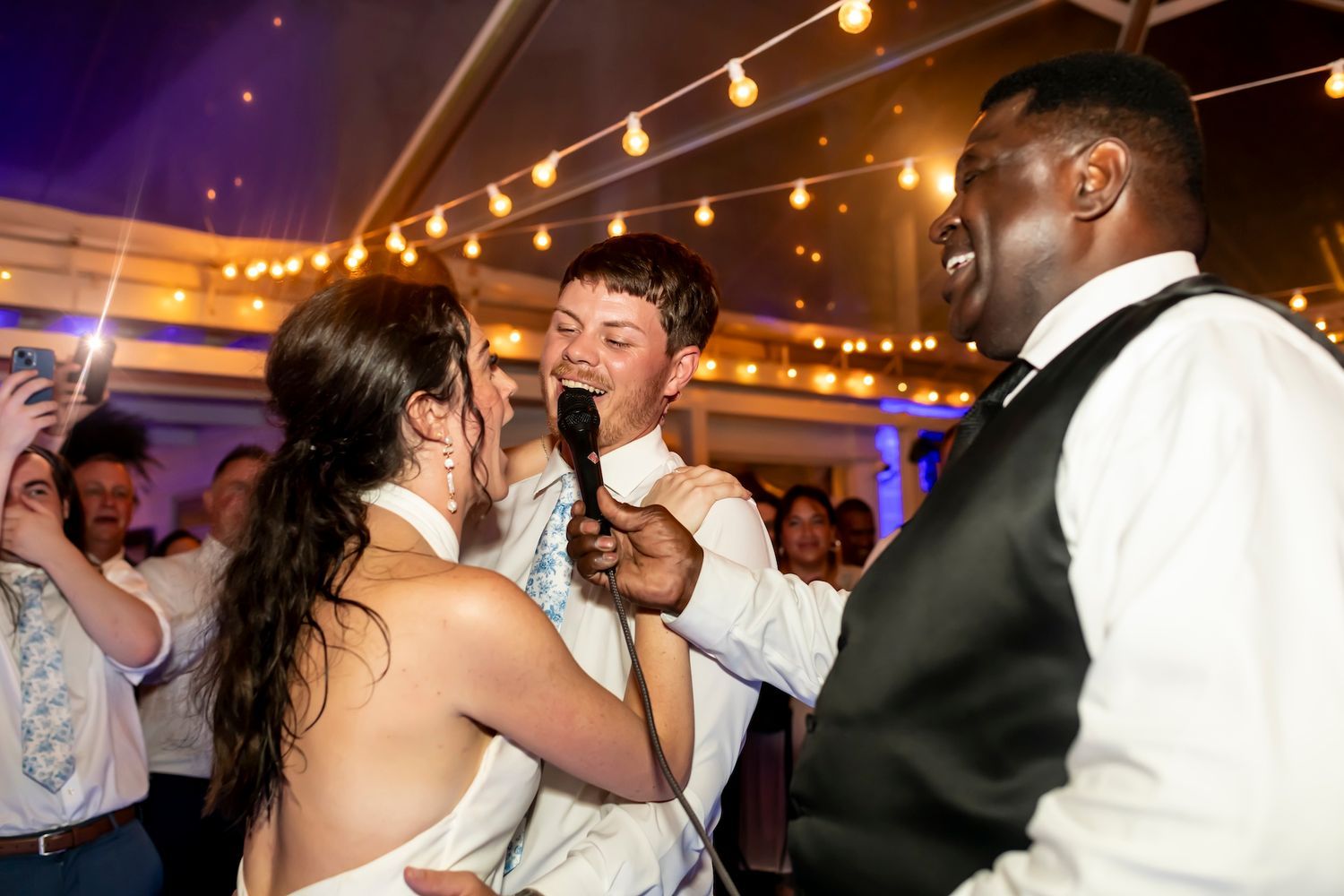 Wedding guests dance and celebrate on the dance floor under warm lighting at an indoor reception venue.