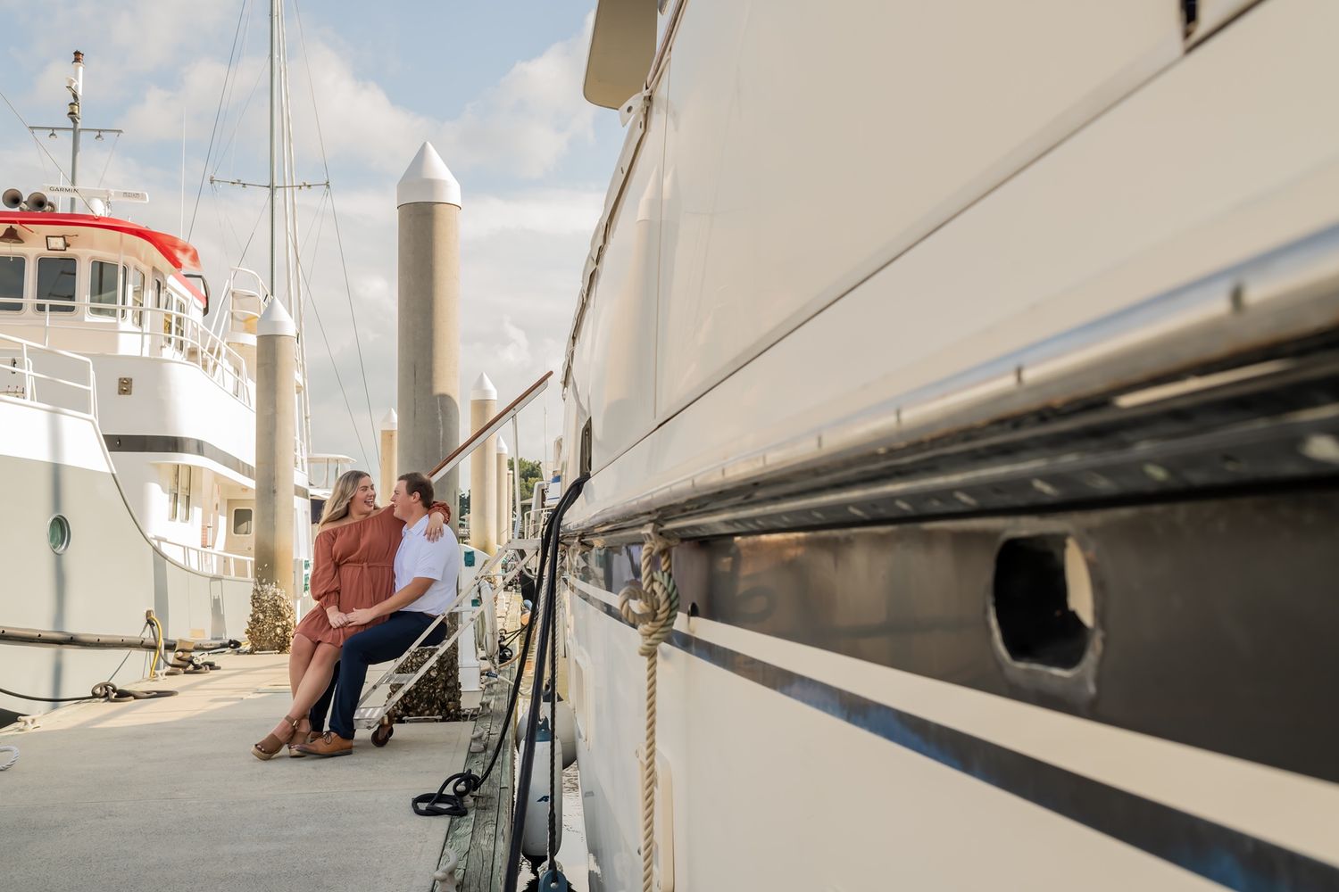 Sailboat masts line the dock at a marina with white vessels and buildings visible in the sunlight.