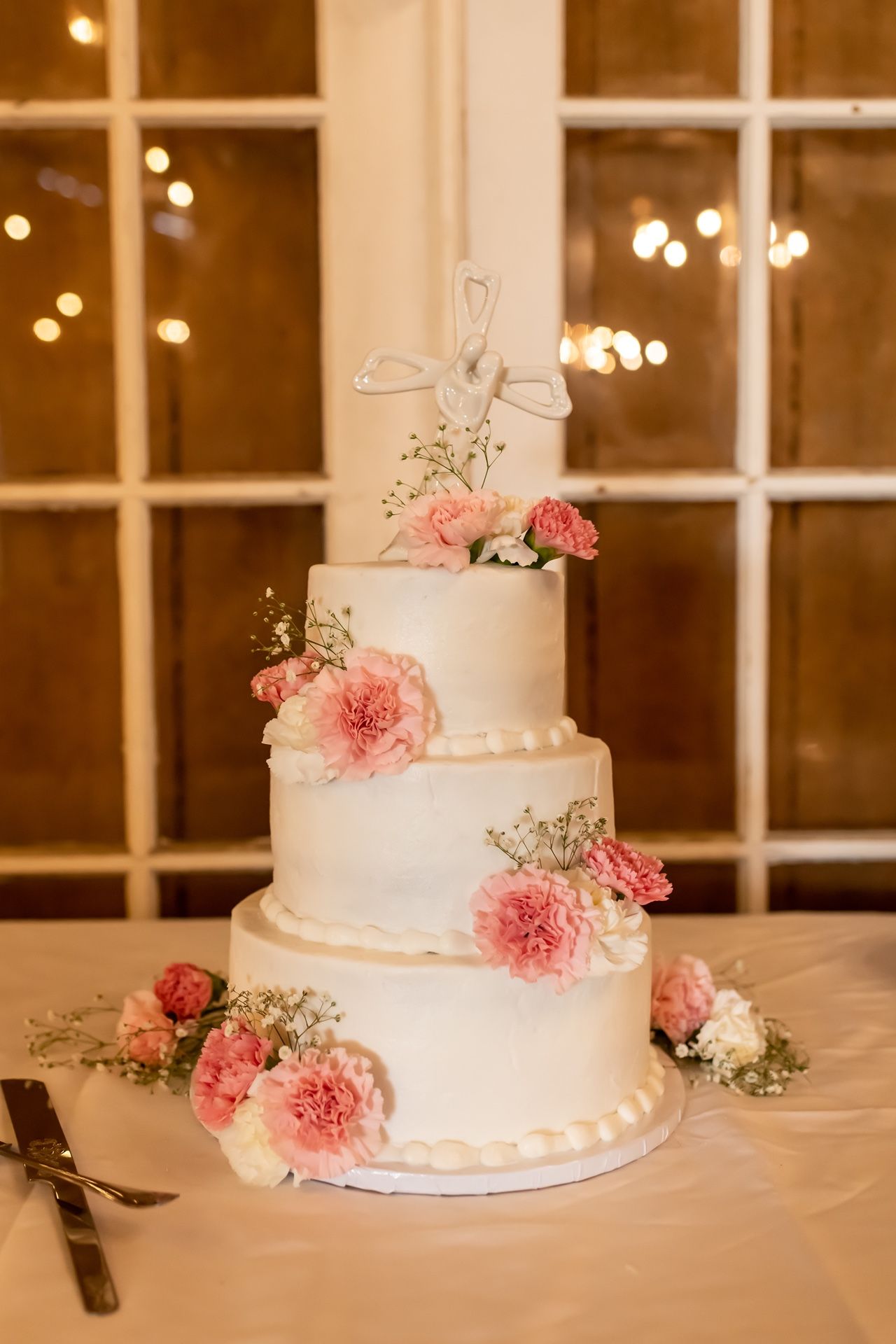 Elegant four-tier white wedding cake decorated with pink garden roses and baby's breath against a warm window backdrop.
