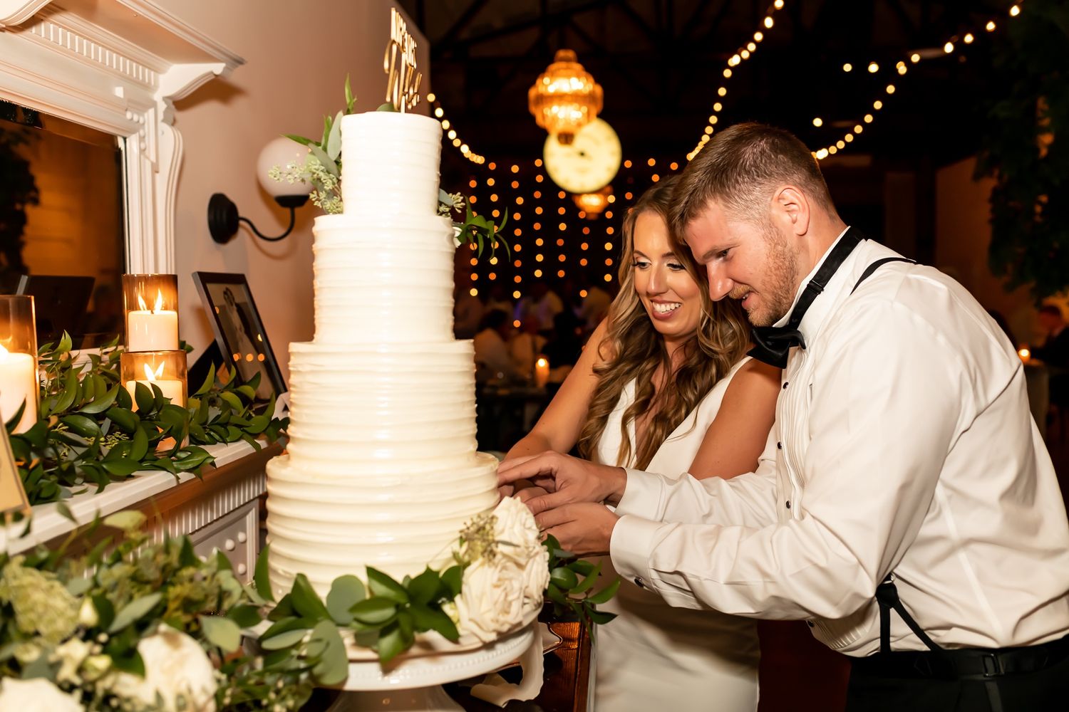 A couple cuts into their elegant white tiered wedding cake at a romantic reception decorated with twinkling lights.