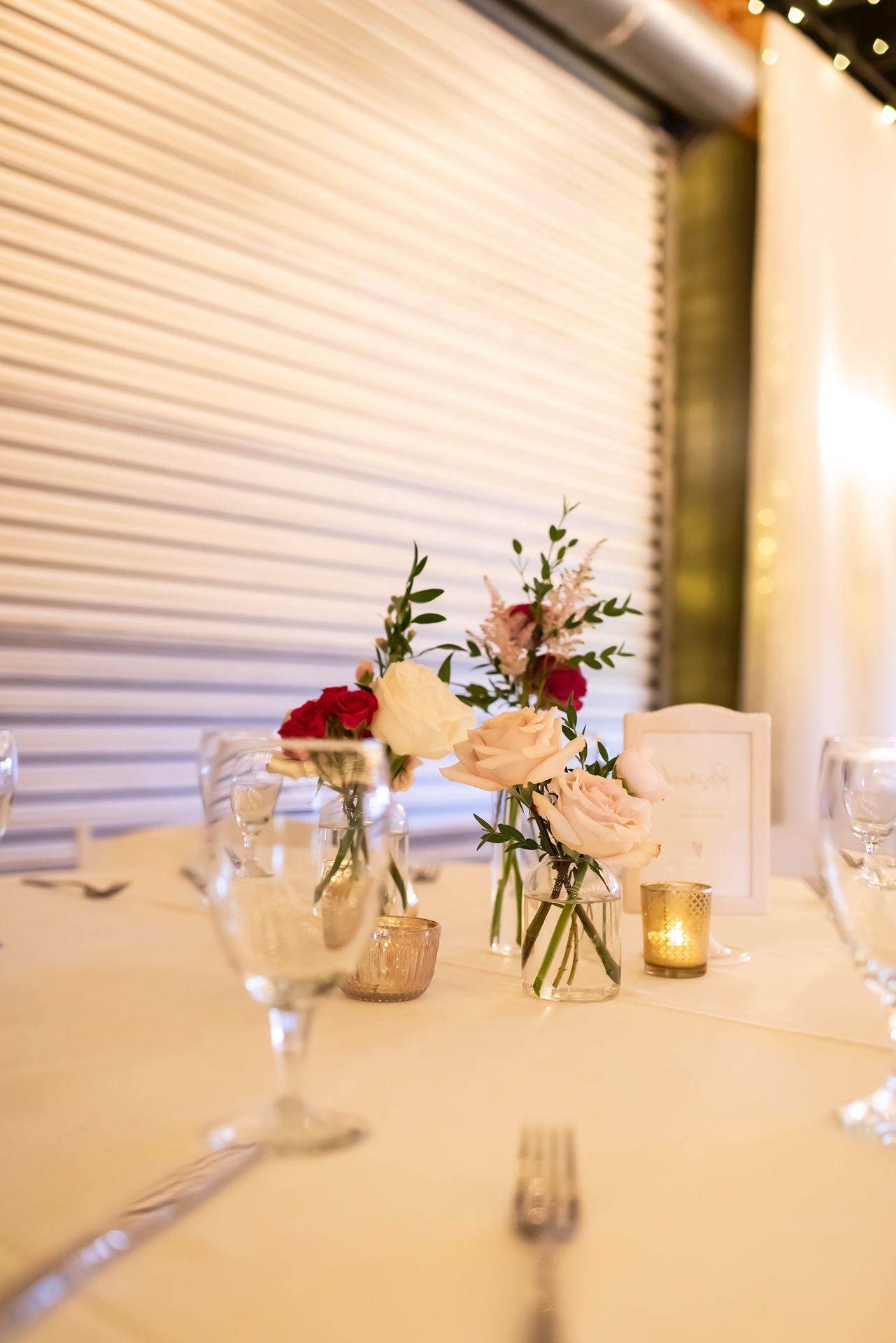 Elegant table setting with small glass vases holding delicate pink and red flowers alongside lit candles at a wedding reception.