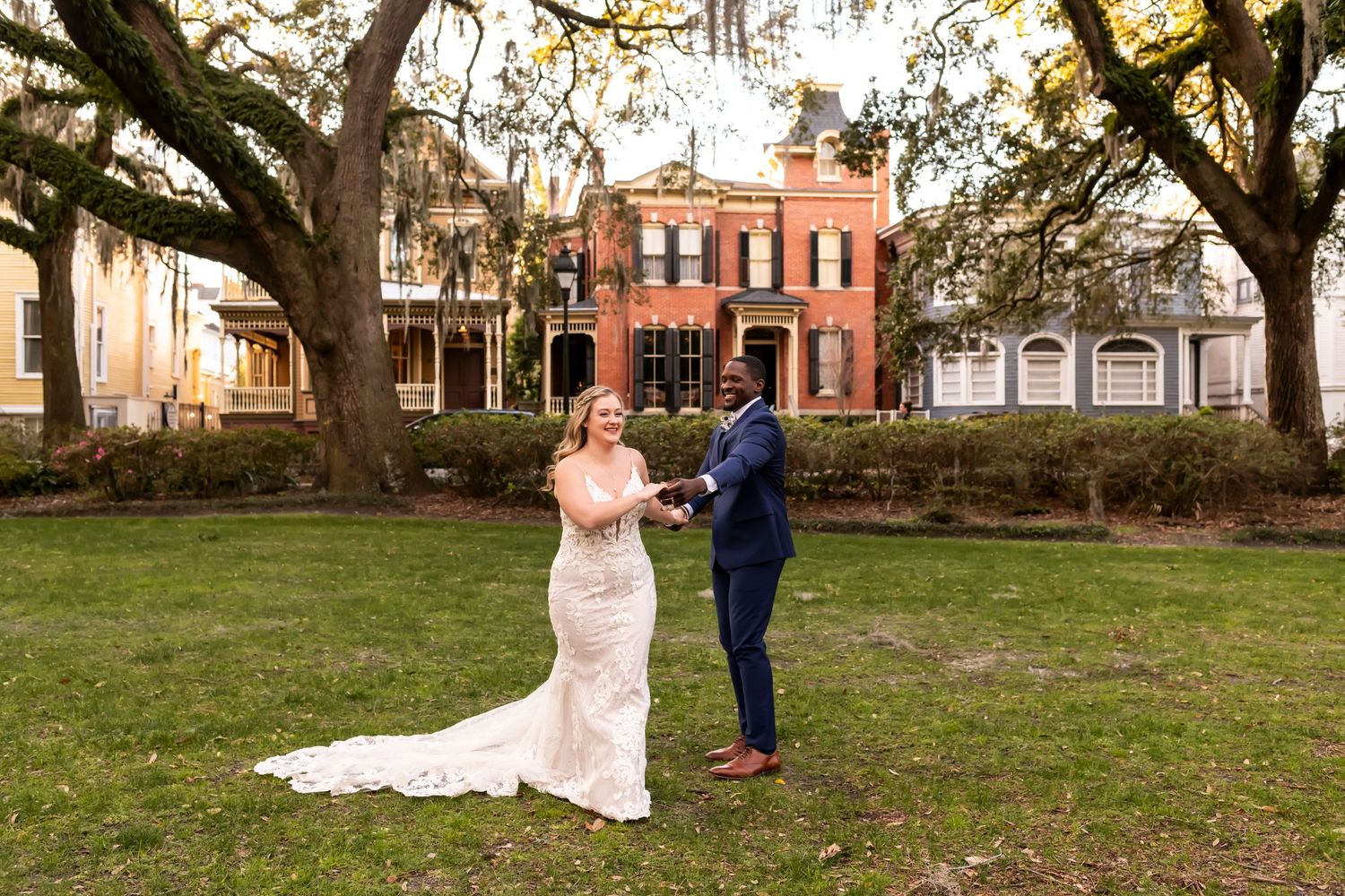 Bride and groom dancing together in courtyard with historic brick building and oak trees in background.