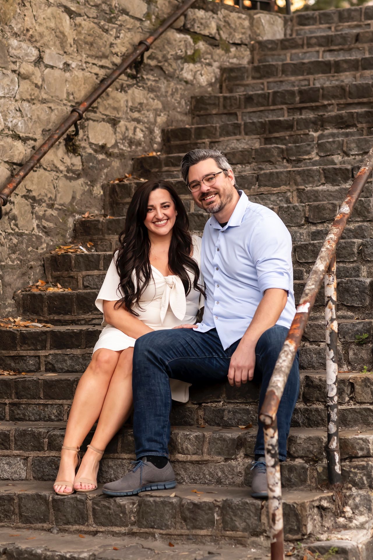 A couple sits together on old stone steps by a rustic wall with wooden poles on a warm evening.