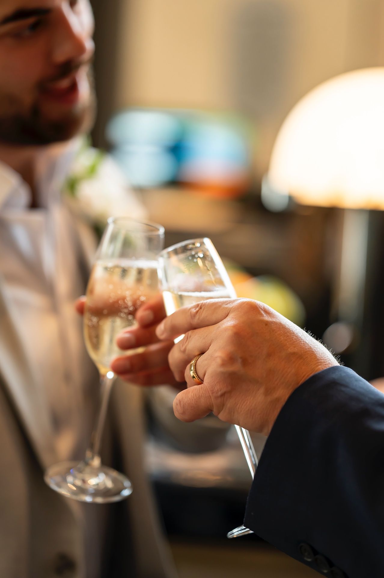 Two hands holding champagne glasses toasting in an elegant setting with a warm, ambient lamp light in background.