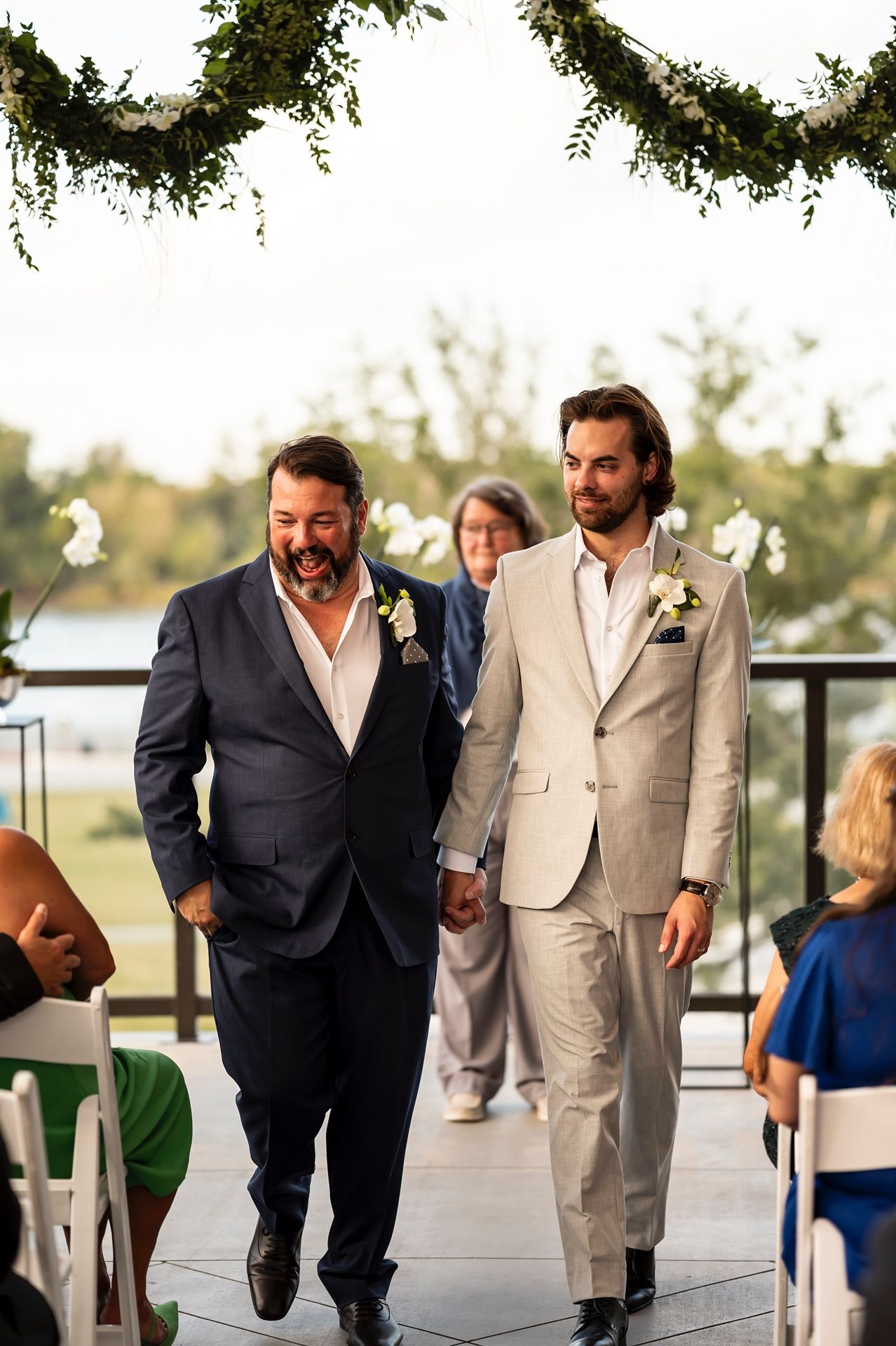 Two grooms walk down the aisle together at an outdoor wedding ceremony under greenery garlands.