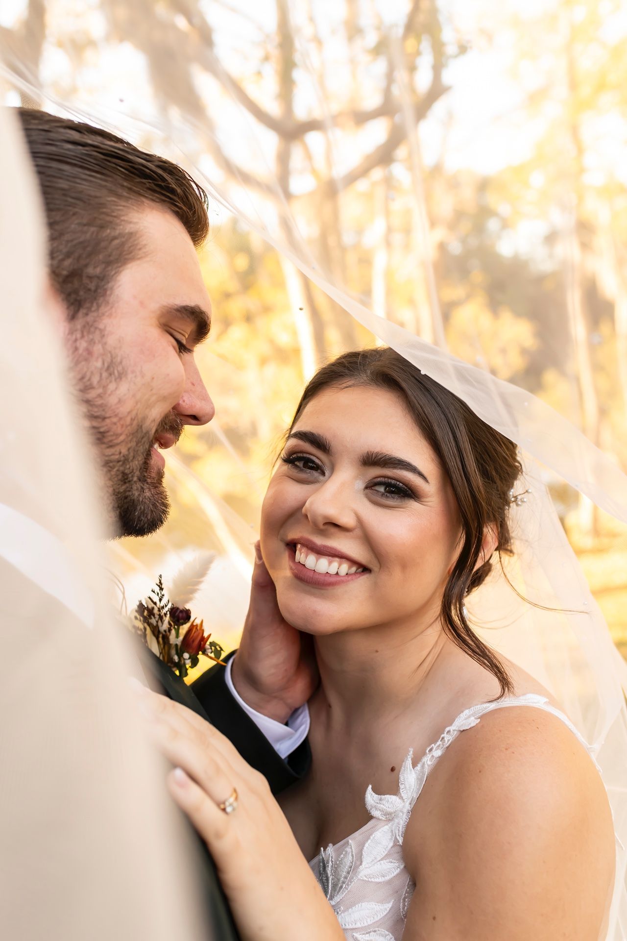 Close up photo of bride and groom sharing an intimate moment under veil with golden sunlight through trees.