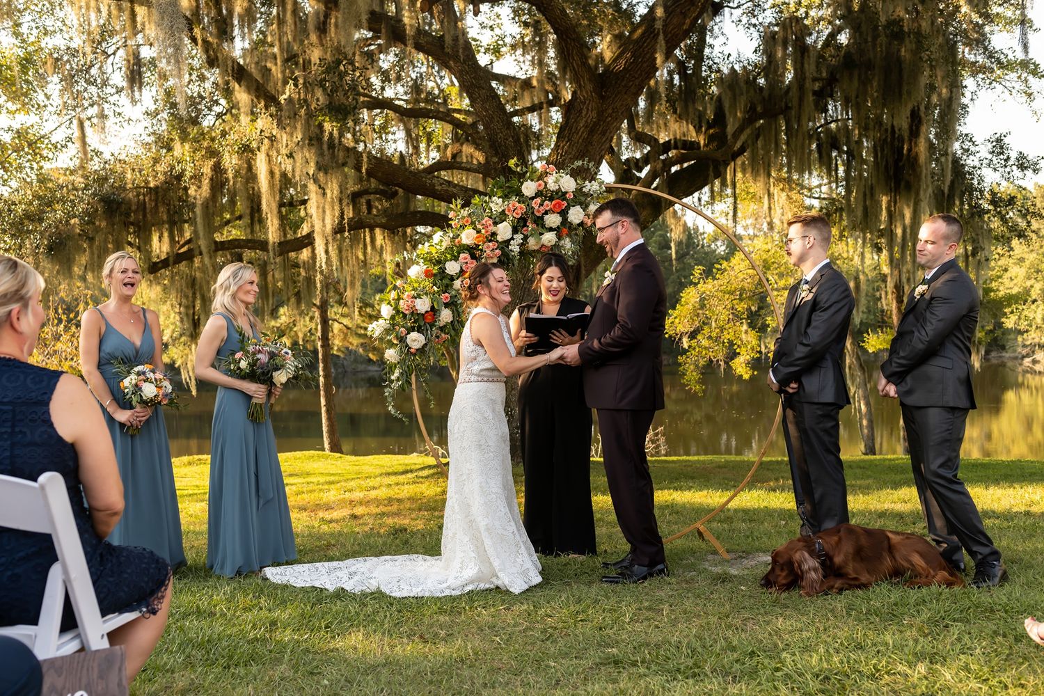 An outdoor wedding ceremony during golden hour features a gathering of people under moss-draped oak trees in a natural setting.