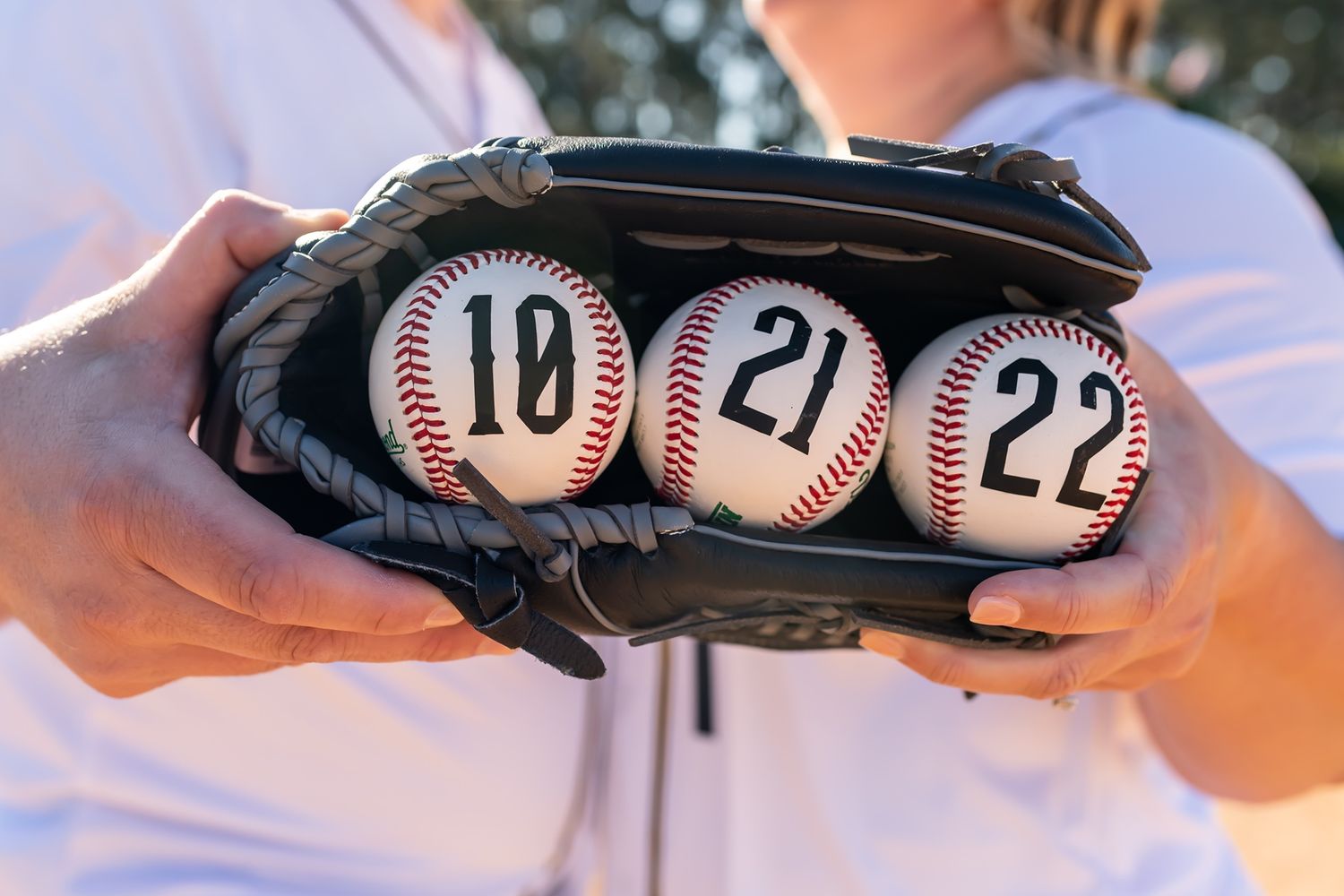 Three baseballs with numbers 18, 21, and 22 sit inside a black baseball glove held in someone's hands.