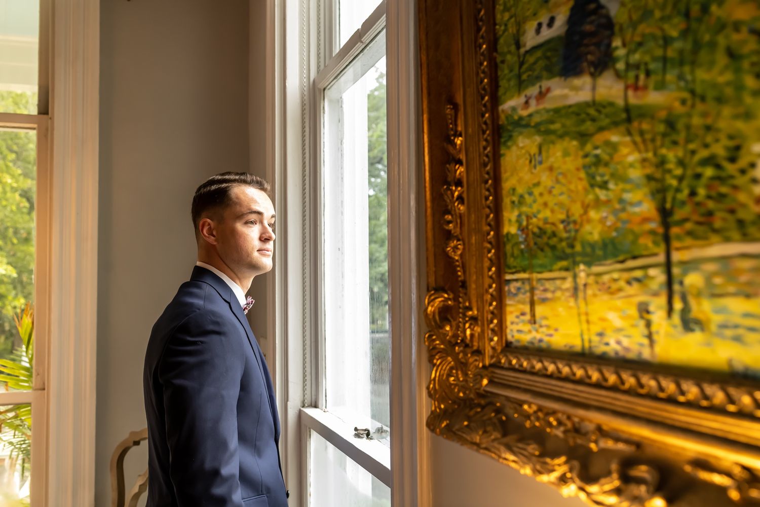 A person in a navy suit stands near large windows in an elegant room with golden-hued lighting and ornate decor.