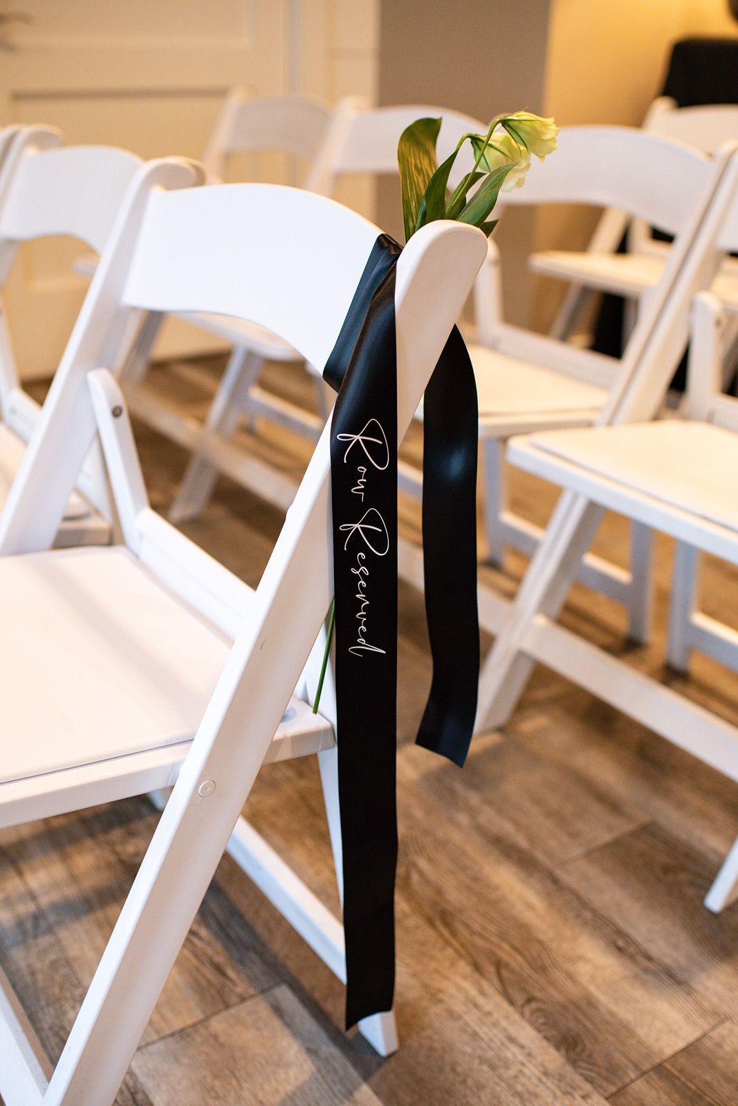 White wooden folding chairs with black sashes tied on the backs line up on a rustic hardwood floor at a wedding venue.