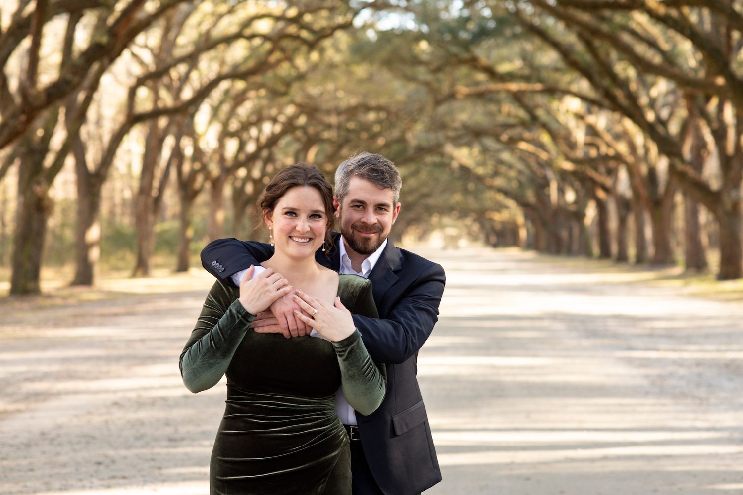 Couple embracing on a tree-lined path, woman in green velvet dress, man in dark suit, smiling outdoors.