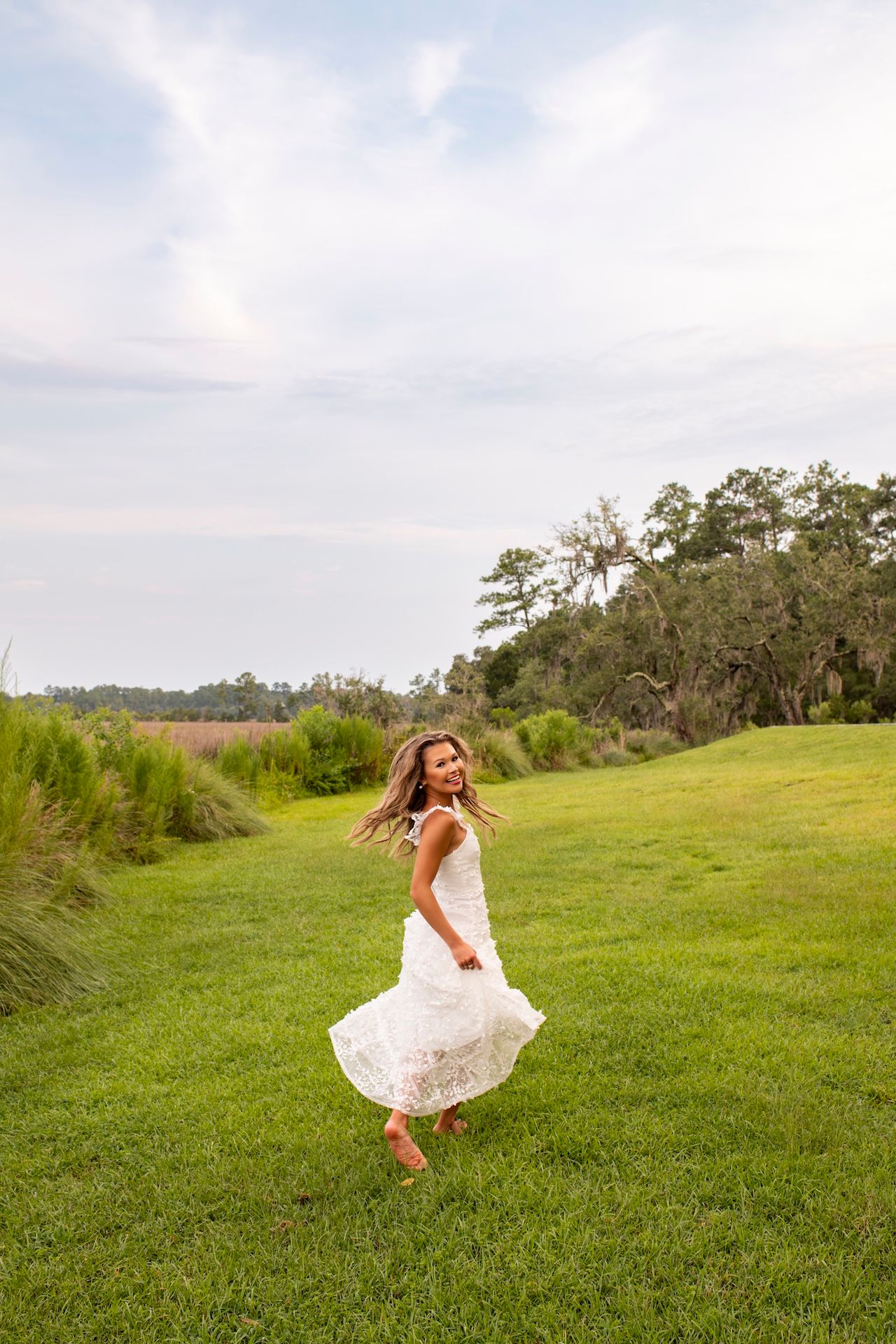 A joyful woman in a white lace dress twirls on a lush green lawn with trees and cloudy sky behind her.