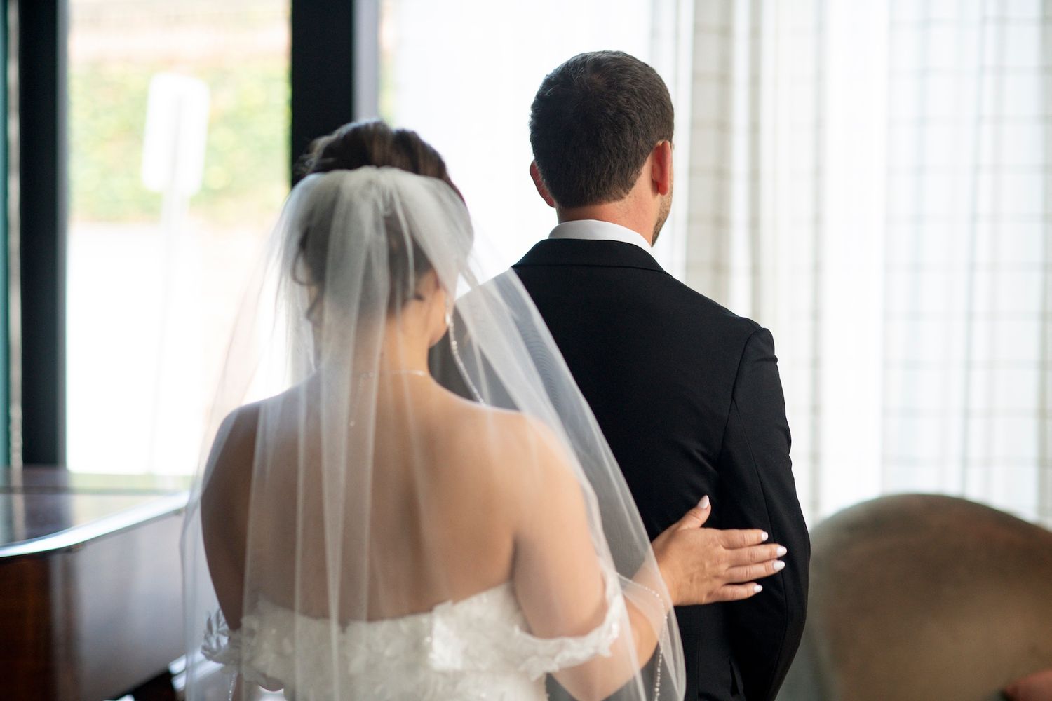 A wedding couple stands together in formal attire with a flowing white veil visible in the sunlight.