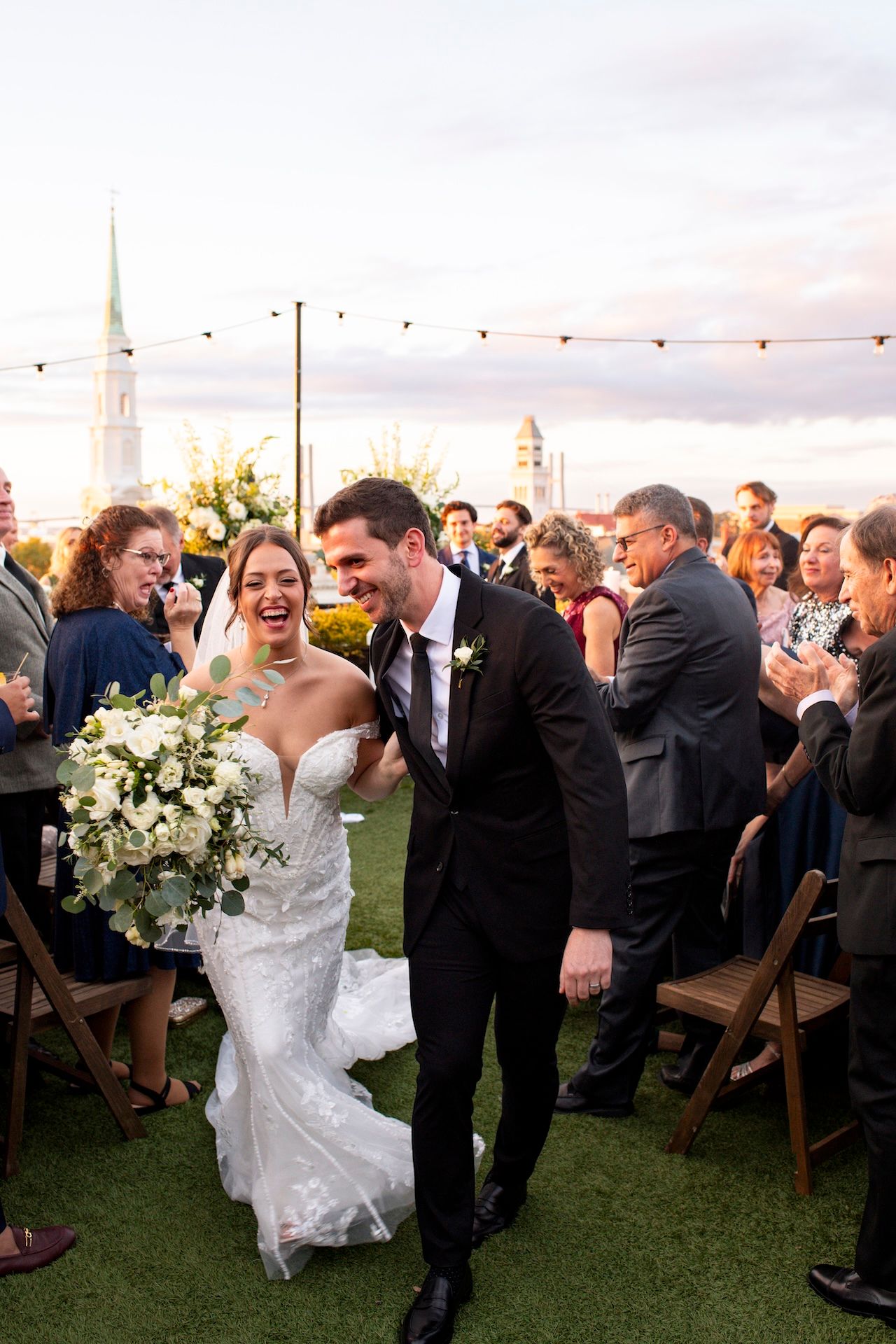 A joyful wedding celebration on a lawn with guests cheering as the newlyweds walk down the aisle at sunset.