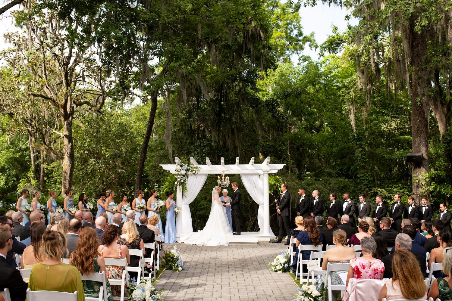 Couple exchanges vows under a white pergola arch surrounded by guests at an outdoor garden wedding ceremony.