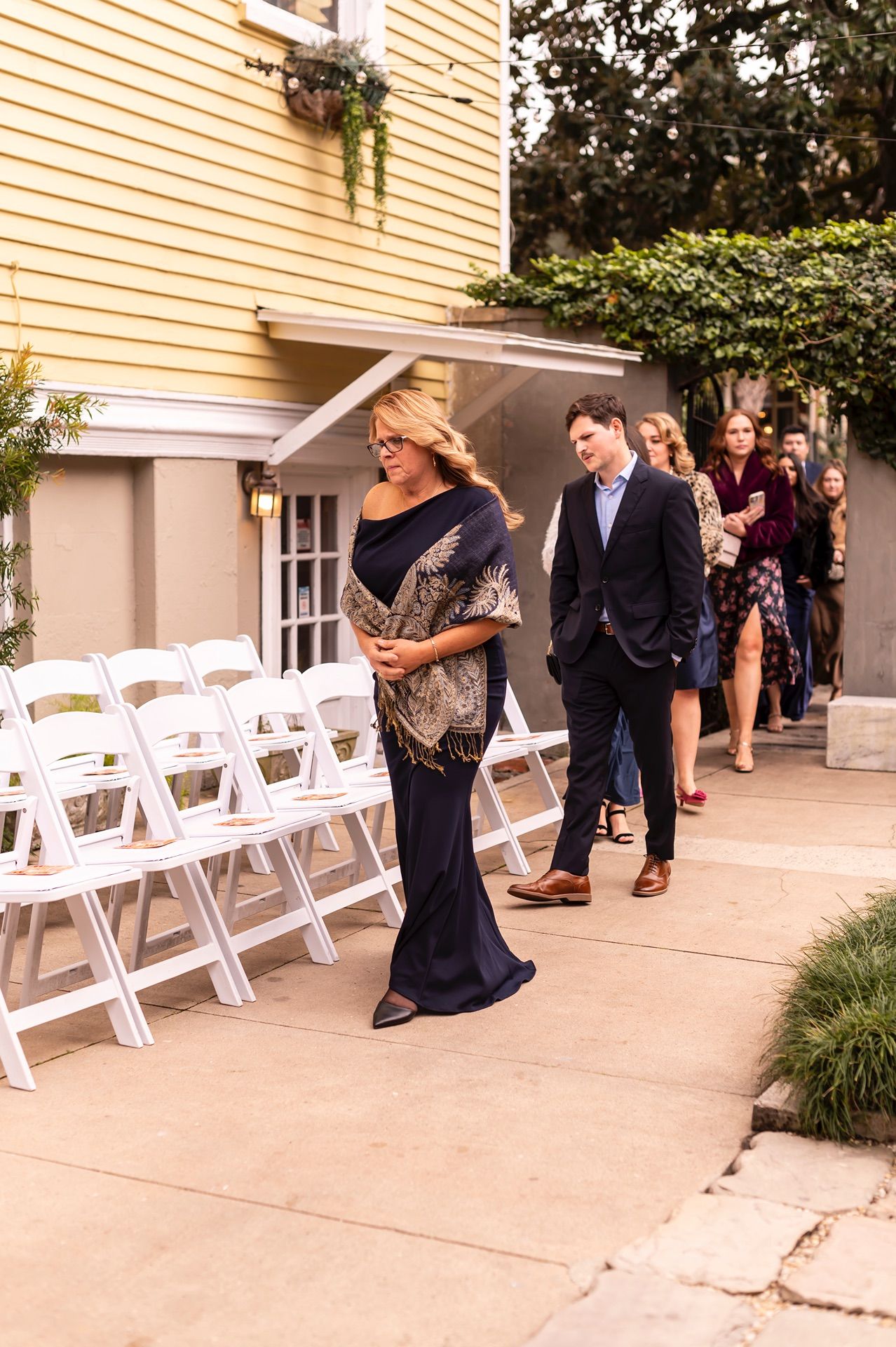 Wedding guests in formal attire walking along a path with white folding chairs lined up for an outdoor ceremony.
