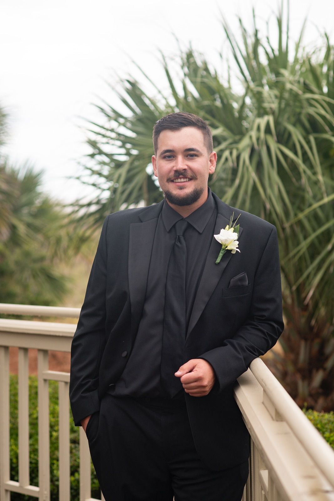 Several men in black formal suits with white boutonnieres stand on a wooden deck with palm plants in the background.