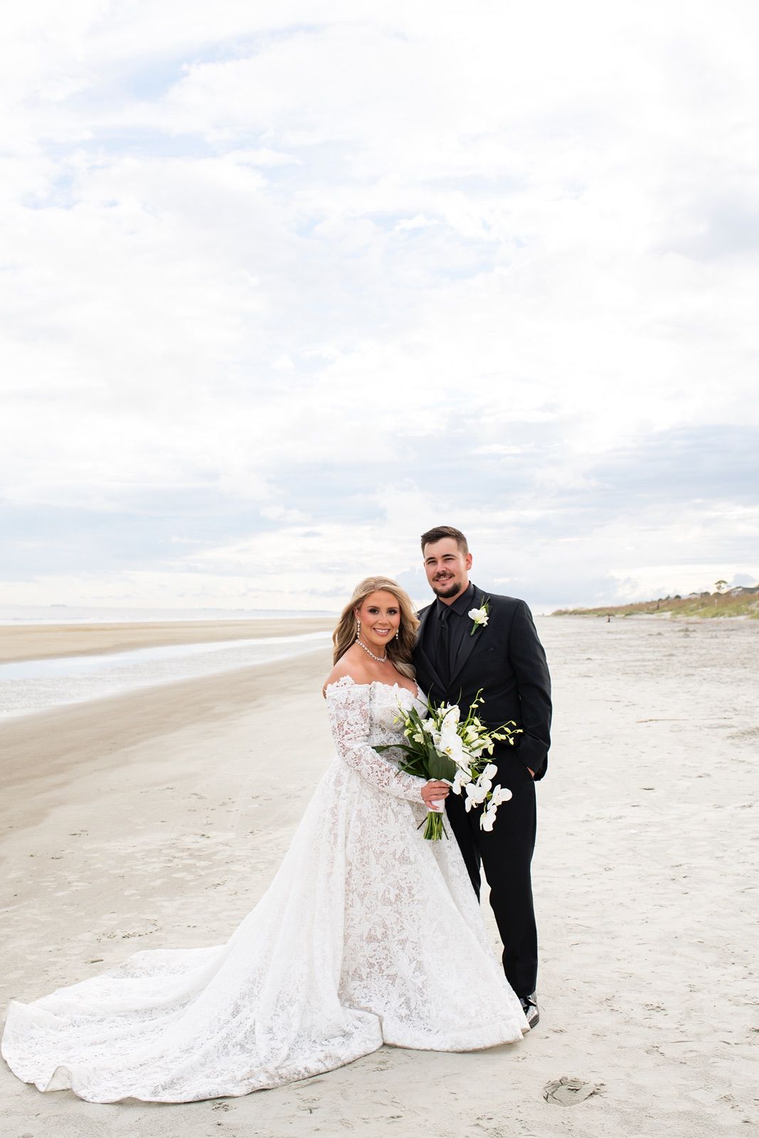 Newlywed couple poses on Hilton Head Island beach with white sand and cloudy skies in the background.