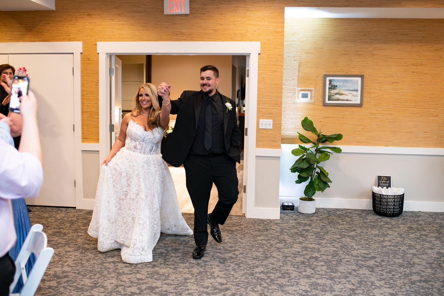 Wedding party members in formal attire walk through a warmly lit hallway with orange walls and carpeted floors.