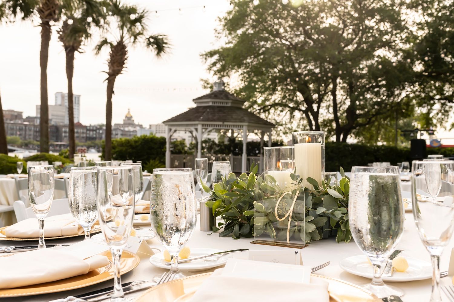 Elegant outdoor wedding reception with white tablecloths, crystal glasses, greenery centerpieces, and a gazebo backdrop at sunset.