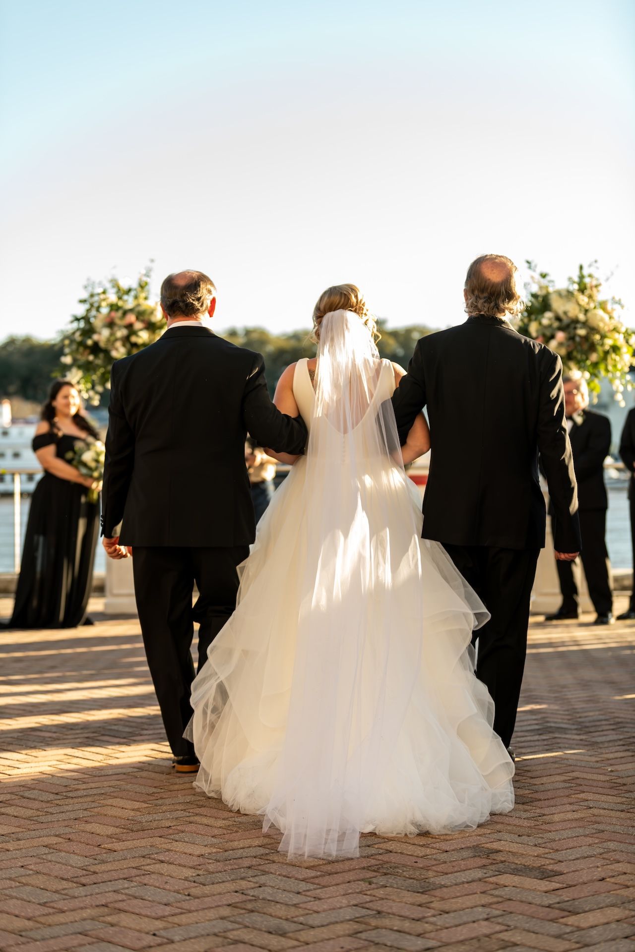 Bride in white gown and veil walks down the aisle flanked by two men in black suits at an outdoor waterfront wedding.