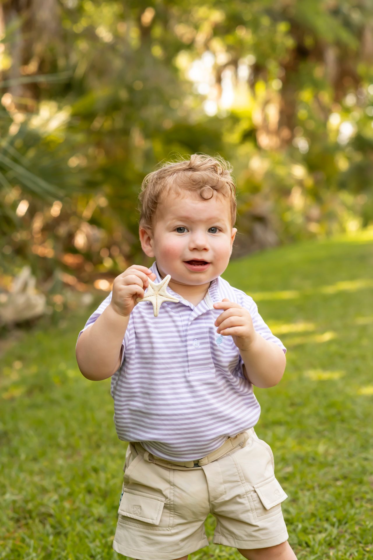 A young child in a striped polo shirt and khaki shorts stands on grass in a sunlit garden setting.