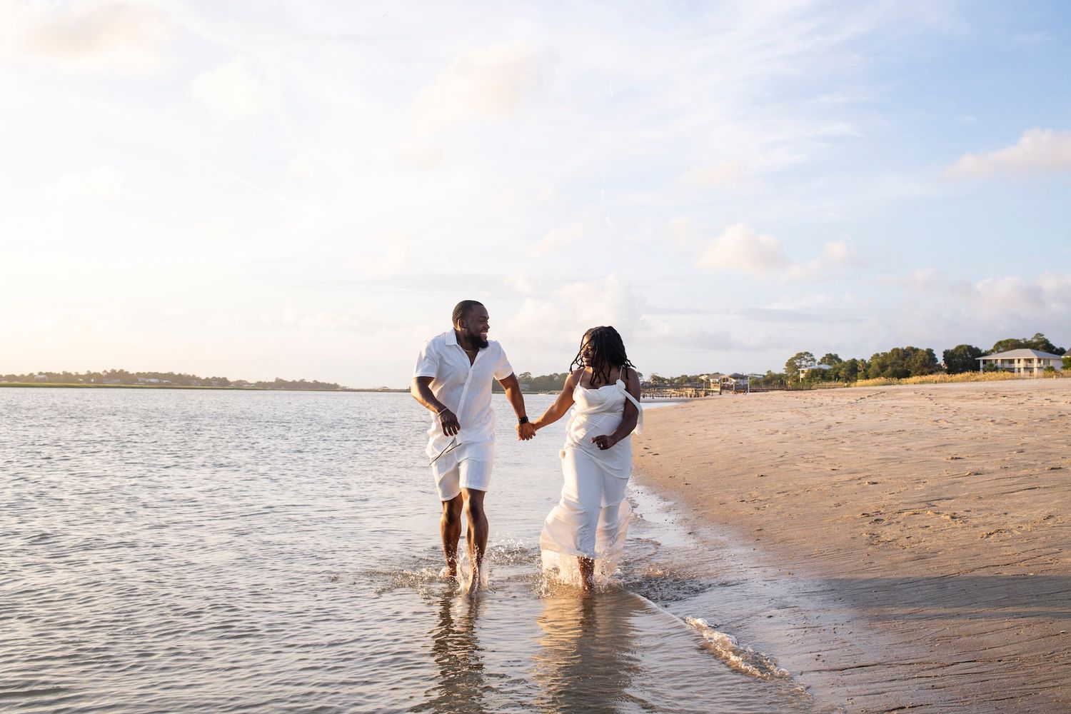 A couple walks along a sandy beach holding hands with ocean waves lapping at their feet during sunset.