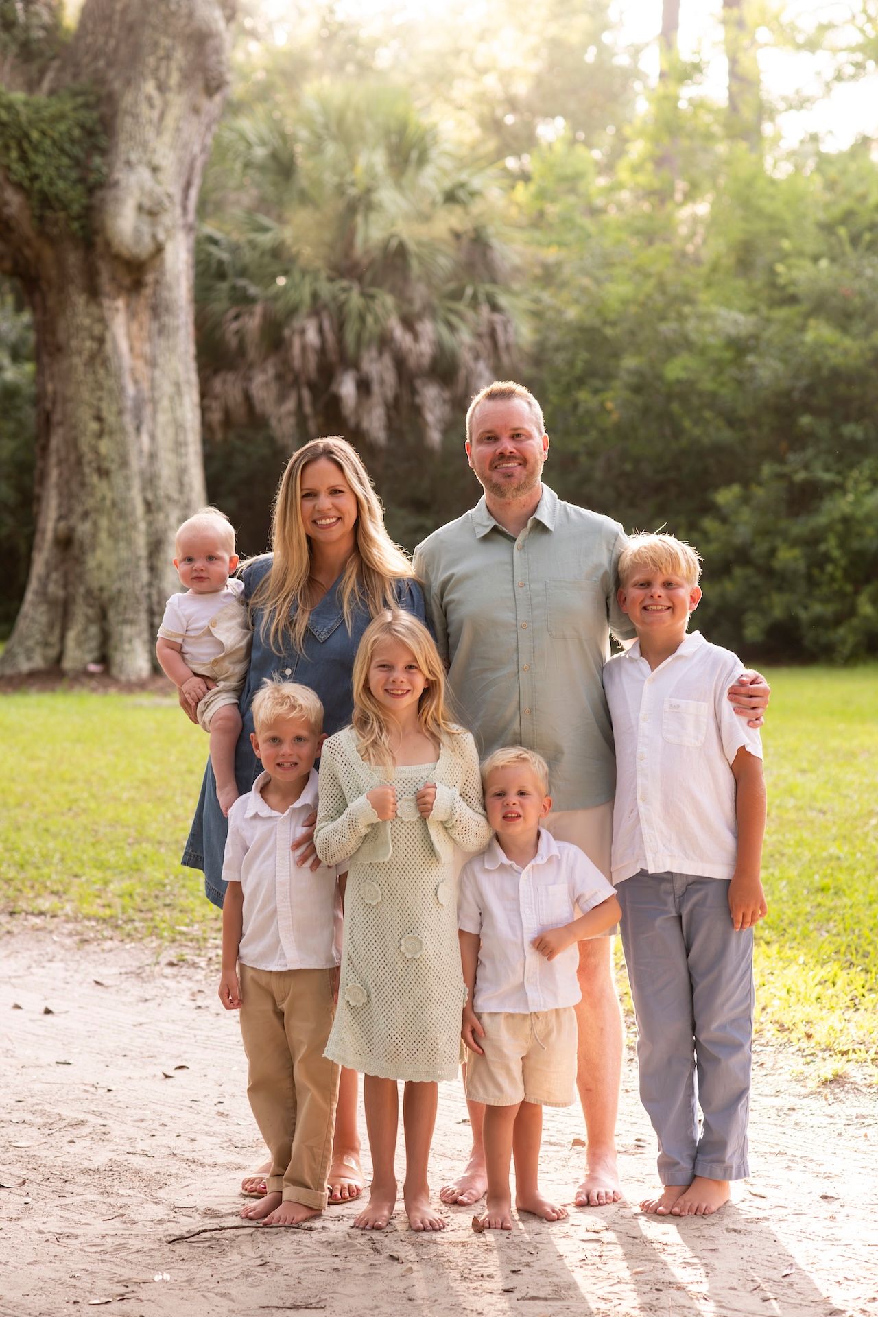 Family of seven posing outdoors on sandy path surrounded by lush green trees in warm golden sunlight.