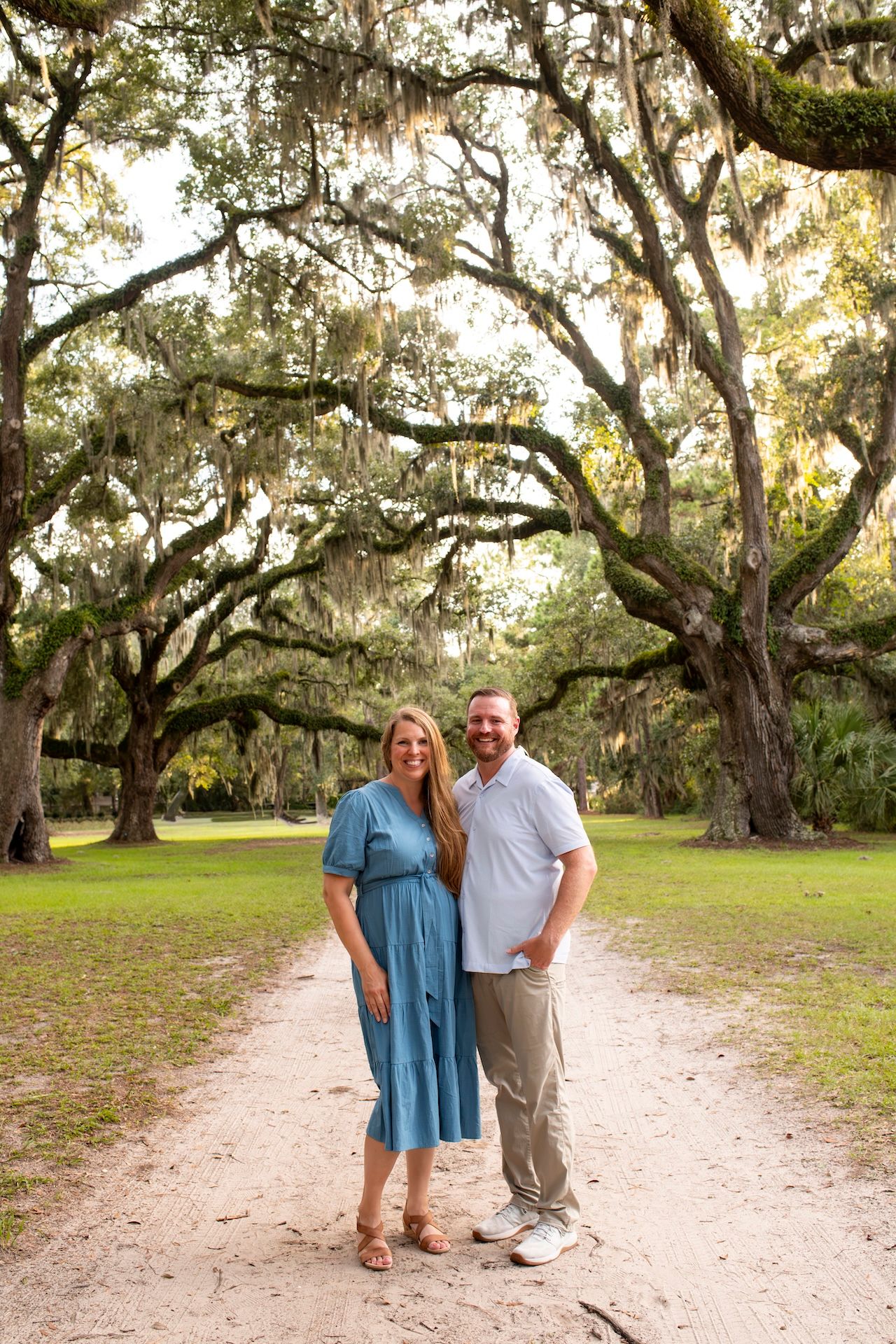 Couple smiling on a sandy path surrounded by large oak trees draped with Spanish moss in a lush park.