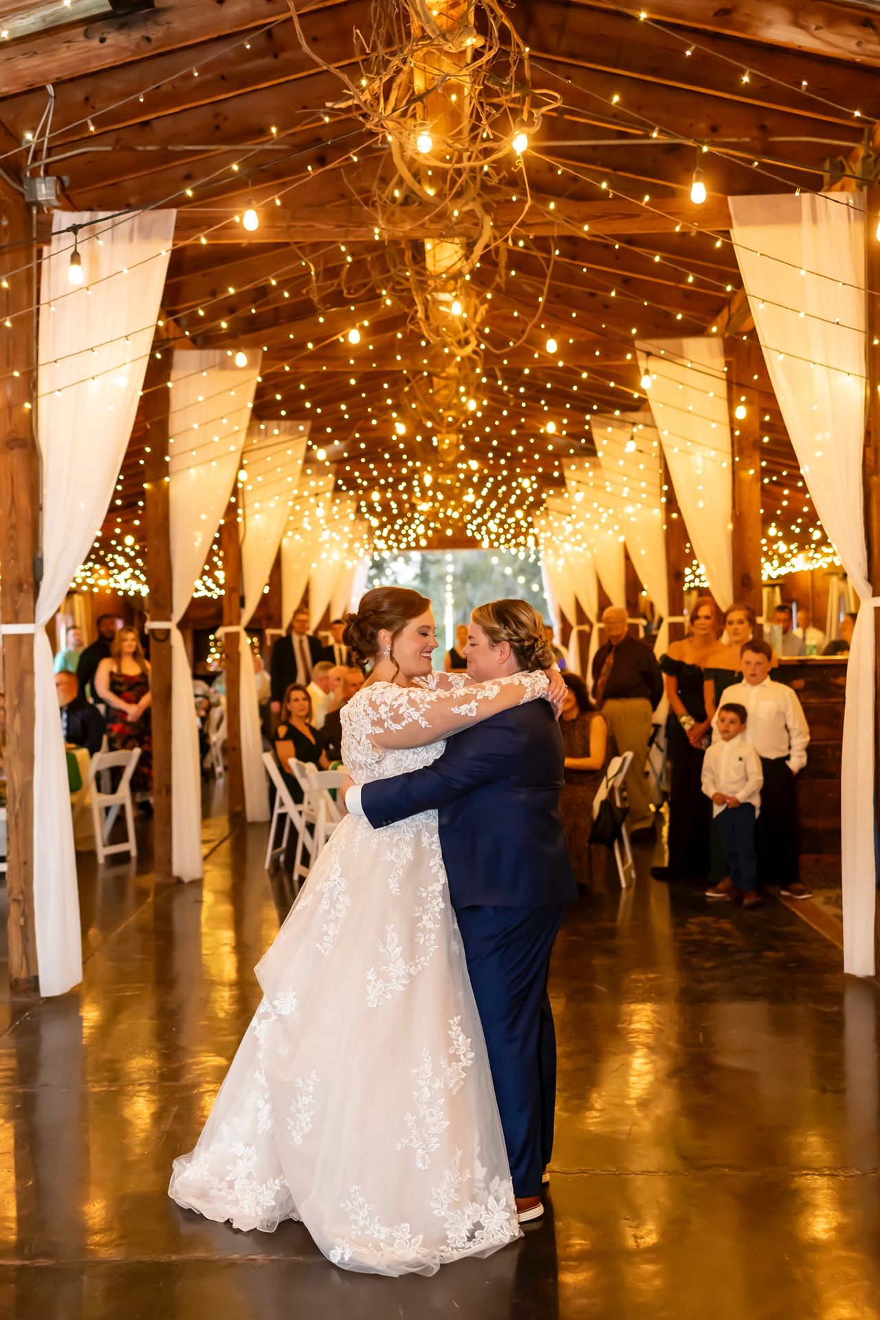 A couple shares their first dance in a rustic barn venue decorated with string lights and white drapes creating a magical atmosphere.