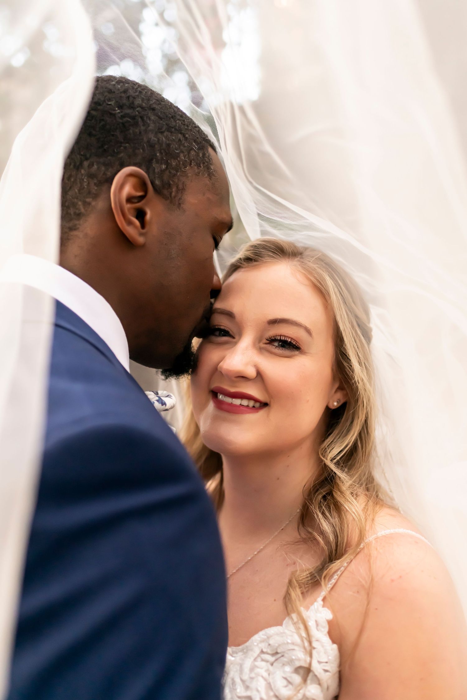 A romantic wedding moment captured under a flowing white veil as a bride smiles joyfully in her lace gown.