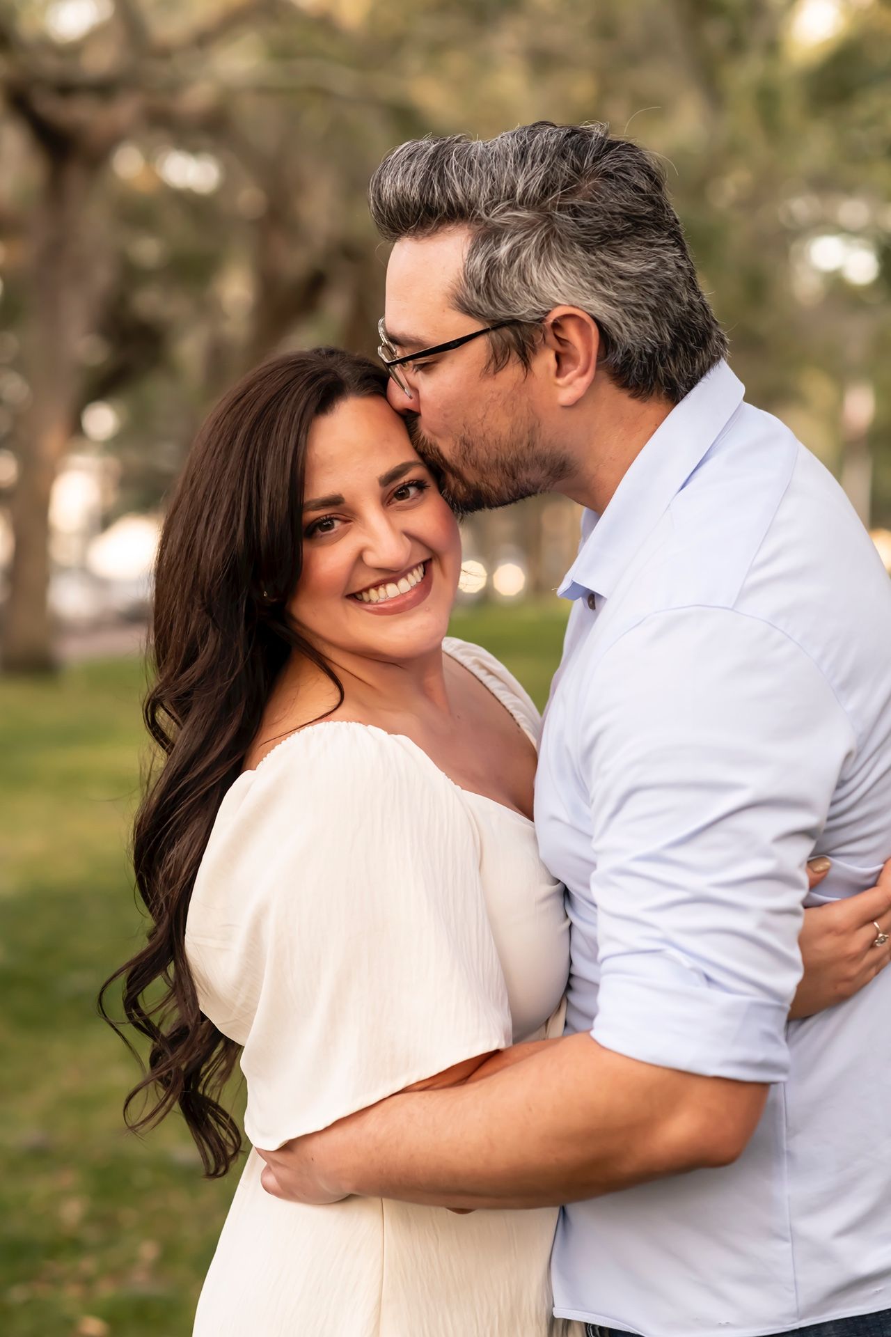Romantic couple embraces outdoors in a park setting during golden hour, wearing white and light colored clothing.