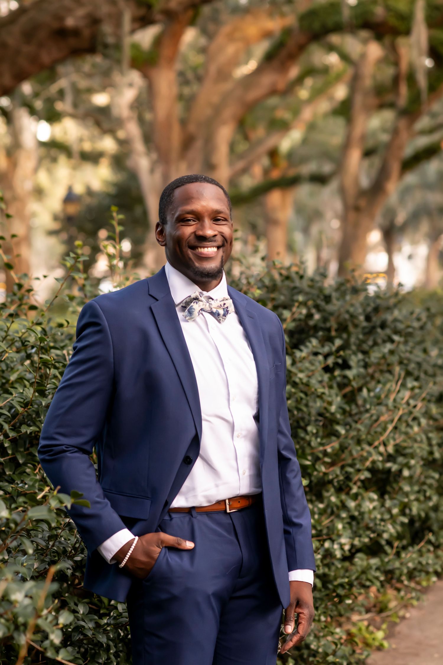 A person in a navy blue suit and floral bow tie smiles warmly while standing in a garden setting with trees in background.