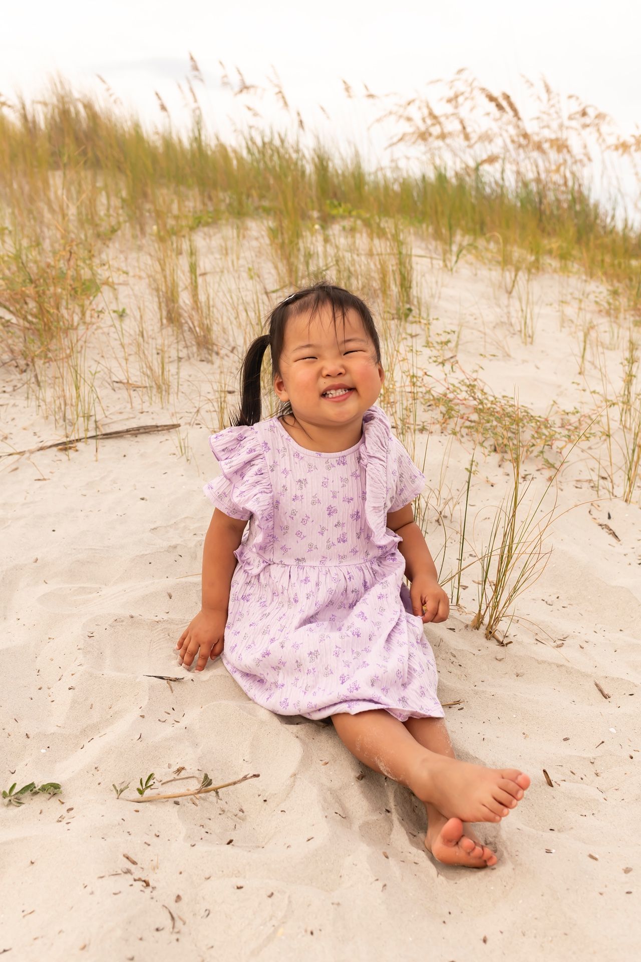 A child in a pink dress laughs while sitting on sandy beach dunes with tall grass behind.