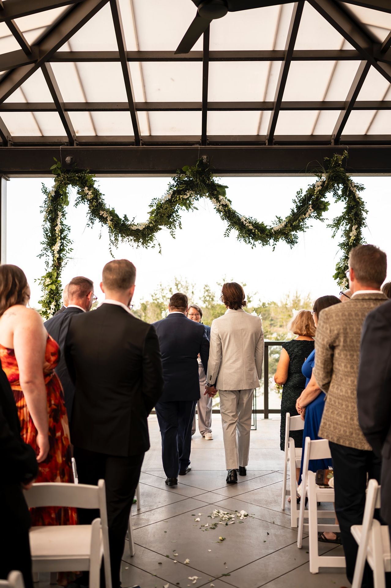 Wedding guests look on during outdoor ceremony under glass atrium with hanging greenery garlands.