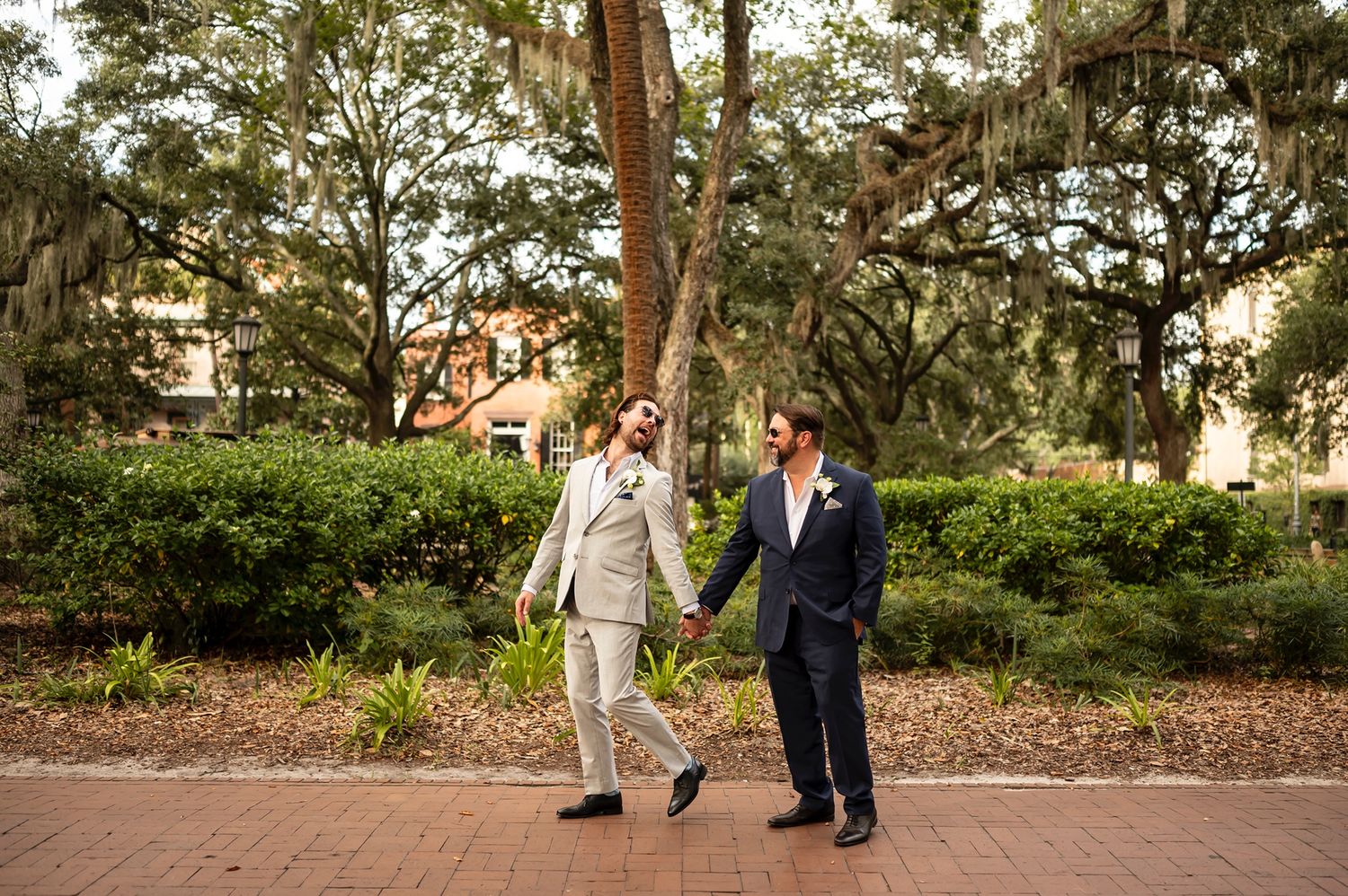 Two grooms taking a romantic walk down a brick path surrounded by lush greenery in a southern garden setting.